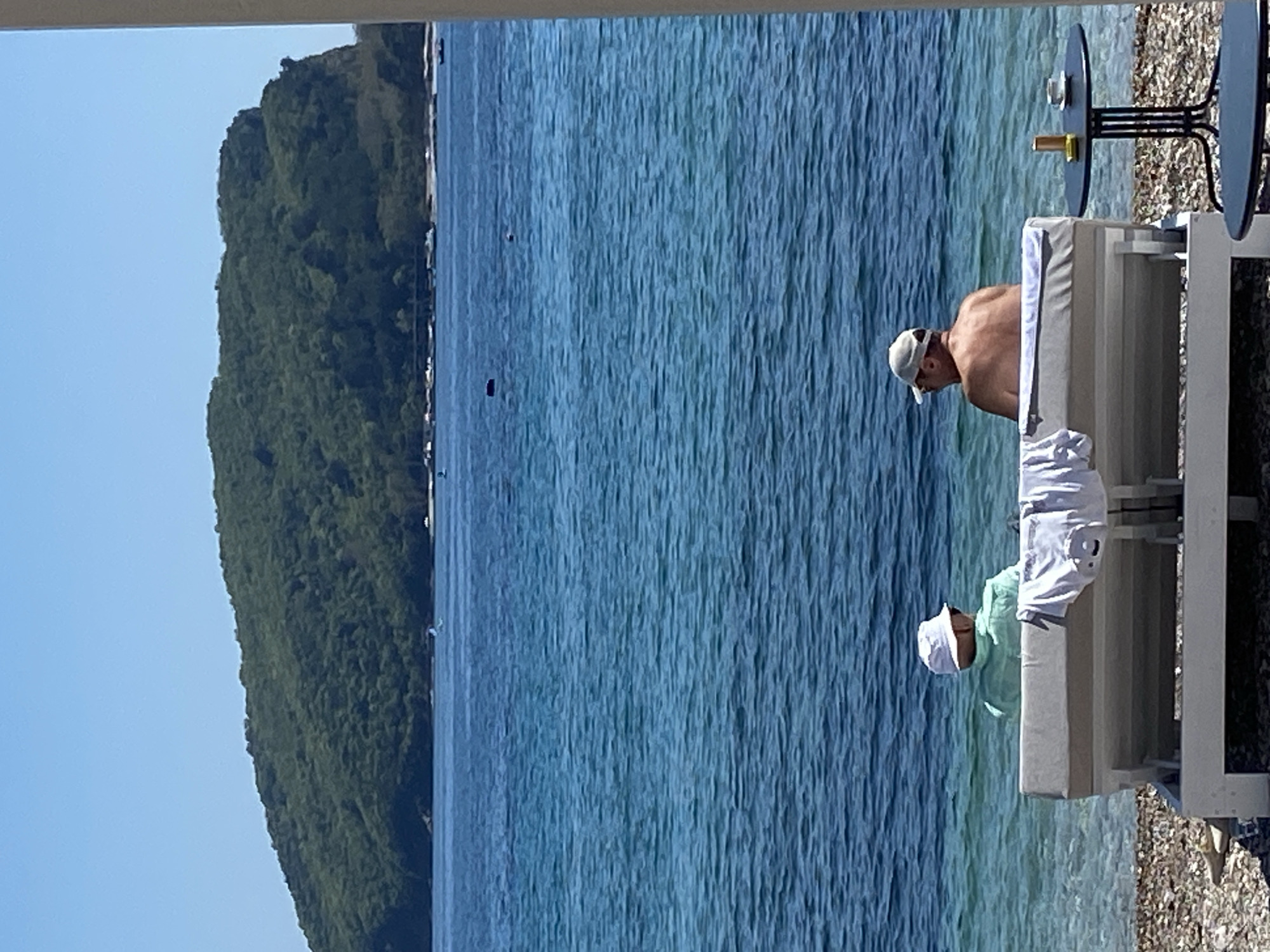 The image shows a coastal scene in Budva, Montenegro. A man with a baseball cap is seated on a beachside lounge chair, situated next to a table. The view encompasses the Adriatic Sea, with a tree-covered mountainous island in the background, and the sky.