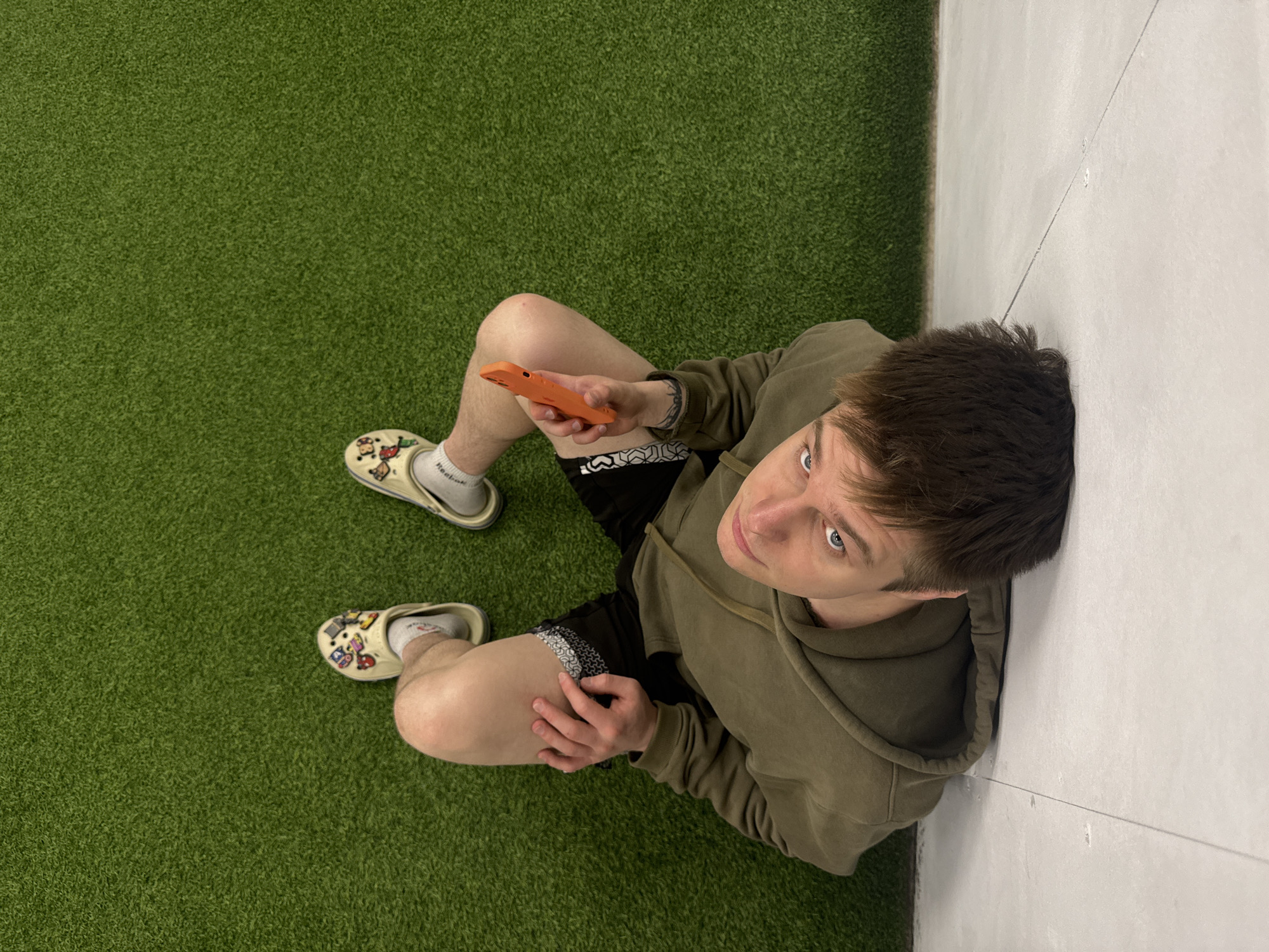 A young man is captured in a high-angle shot, sitting on vibrant green artificial turf and leaning against a light-colored, possibly concrete, wall. He is wearing an olive green hooded sweatshirt and black shorts with a patterned waistband visible. On his feet, he has white socks, one clearly displaying "Reebok," and light-colored clogs adorned with various colorful pins or charms. In his left hand, he holds a bright orange smartphone. He looks directly up towards the camera with a neutral and slightly curious expression. The wall behind him shows subtle horizontal lines. The scene suggests an indoor or enclosed recreational area. The location context for this image is Chaikovsky, Russia.