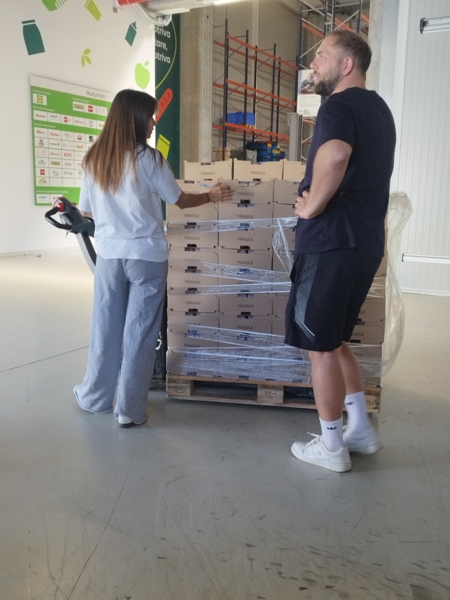 Two individuals, a woman and a man, are standing in a warehouse in Cernica, Romania. The woman, wearing a white t-shirt and grey pants, is operating a pallet jack, positioned to the left of a stack of boxes wrapped in plastic wrap. The man, wearing a dark t-shirt and dark shorts, stands to the right of the boxes, with his hands on his hips. The boxes are labeled "FRAGILE". The warehouse has tall shelving units in the background.