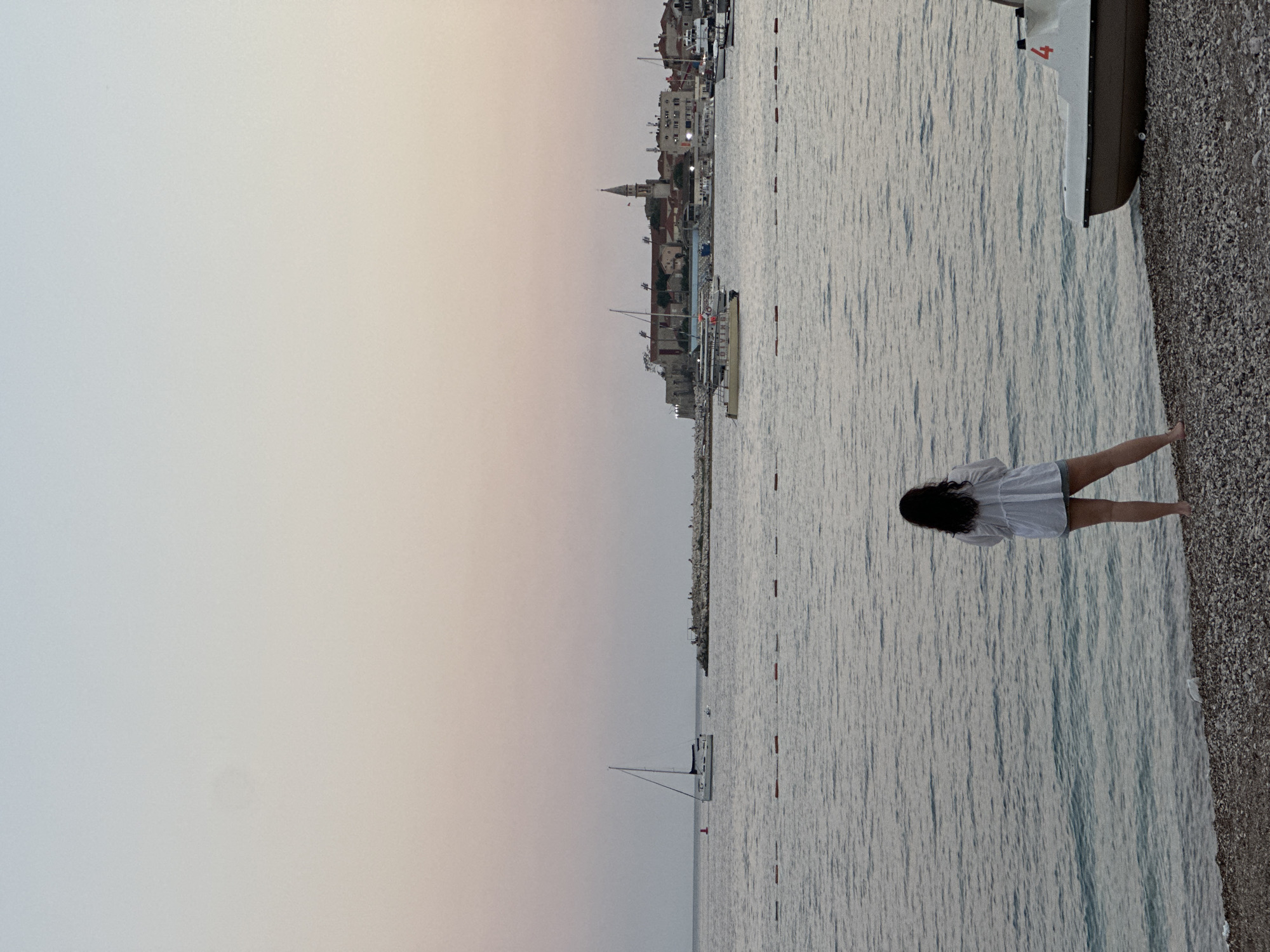 The image depicts a serene sunset scene on the coast of Budva, Montenegro. In the foreground, a person stands on a pebble beach, facing away from the camera, looking out at the calm sea. The person is wearing a white, loose-fitting shirt and shorts. To the right, a small pedal boat is partially visible on the shore. The sea stretches towards a distant peninsula where the historic Old Town of Budva is situated. The town features red-tiled roofs, stone buildings, and a prominent church steeple. Several boats, including a sailboat and smaller motorboats, are scattered across the water, some near the shore and others closer to the Old Town. The sky displays a gradient of soft pastel colors, transitioning from pale blue at the top to a warm pinkish-orange near the horizon, characteristic of a sunset. Floating buoys create a line across the water in the mid-ground. The overall atmosphere is peaceful and tranquil.
