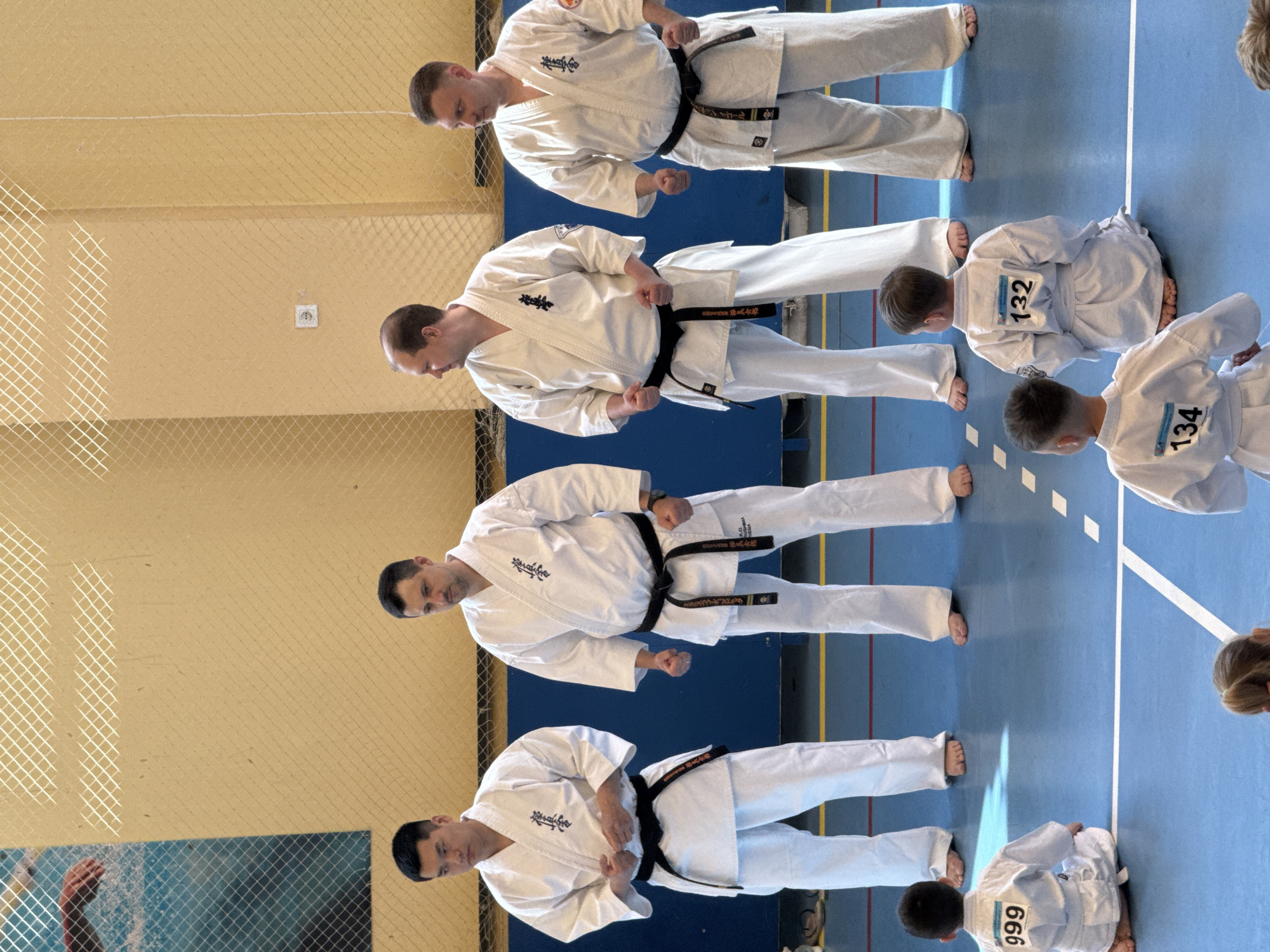 The image depicts a karate competition in Yekaterinburg, Russia. Four adult men wearing white karate uniforms with black belts stand in a line, performing a synchronized stance. The men are barefoot and facing slightly downward. Adjacent to them, several children in similar white uniforms with competition numbers affixed to their backs are kneeling on the blue floor. The backdrop includes a beige wall with a white electrical outlet and a chain-link fence. The floor has white and yellow lines, indicating the boundaries of the competition area. The overall setting suggests an indoor gymnasium or sports hall.