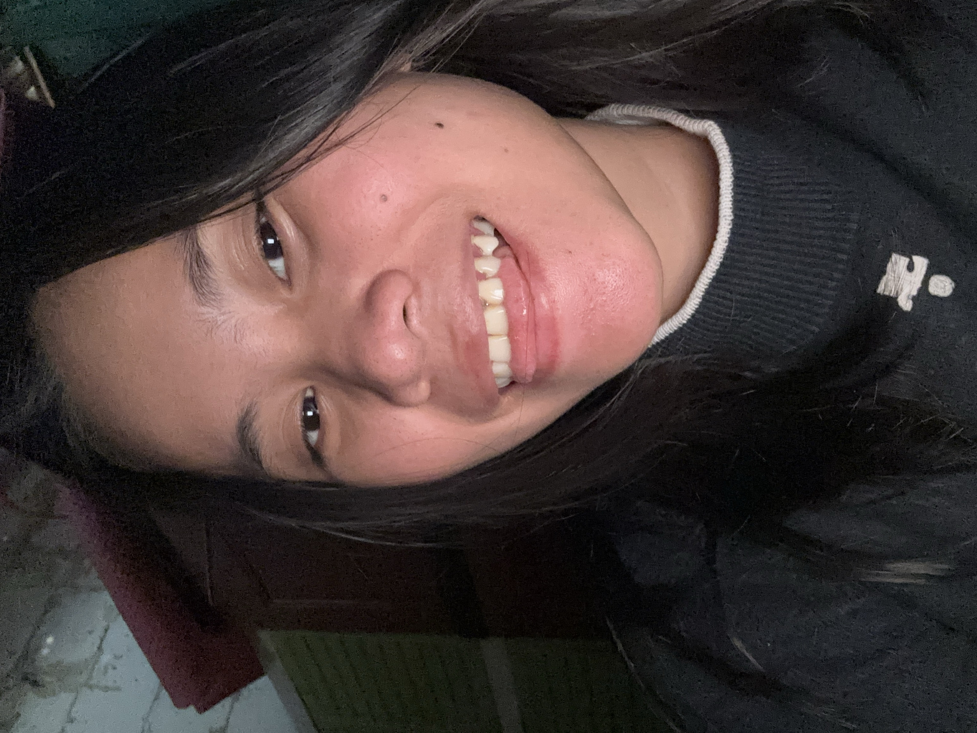 A young woman with long, dark hair is captured in a close-up, selfie-style portrait. She is wearing a black sweater with a light-colored trim around the neckline, and a white graphic element is visible on the lower part of the sweater. Her mouth is slightly open, with her tongue visible, and she appears to be smiling or making a playful expression. The background is dimly lit and appears to be an indoor setting, with hints of textured walls and what might be furniture or shelves. The lighting is somewhat harsh, casting shadows and highlighting the woman's facial features. The image was taken in East Jakarta, Indonesia.