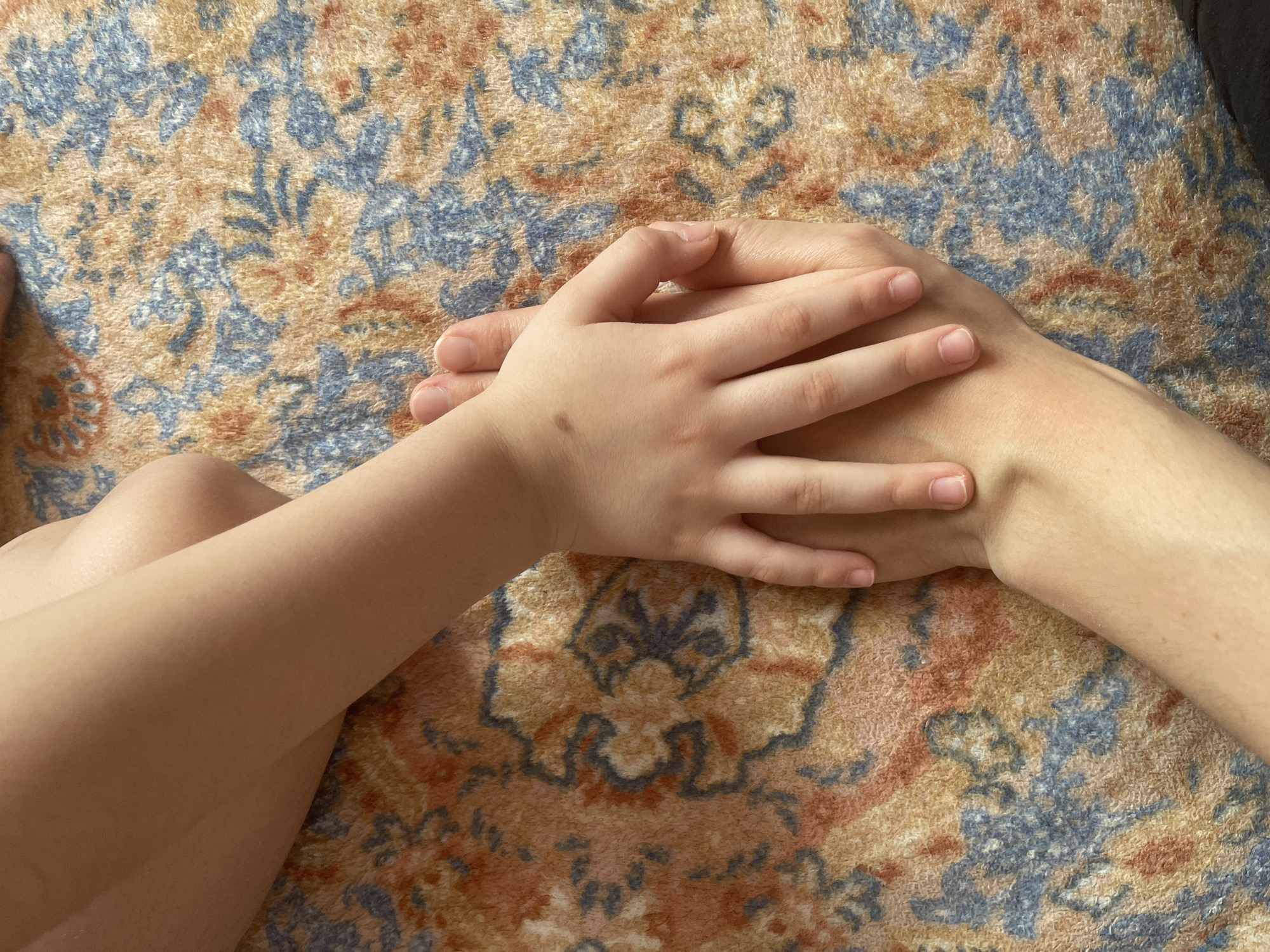 A child's hand rests upon an adult's hand against a patterned rug. The rug features a repeating motif in shades of blue, orange, and tan. Part of a child's leg is visible in the lower left corner of the image. The setting appears to be indoors. There is no information that this image was taken in Yessentukskaya, Russia.