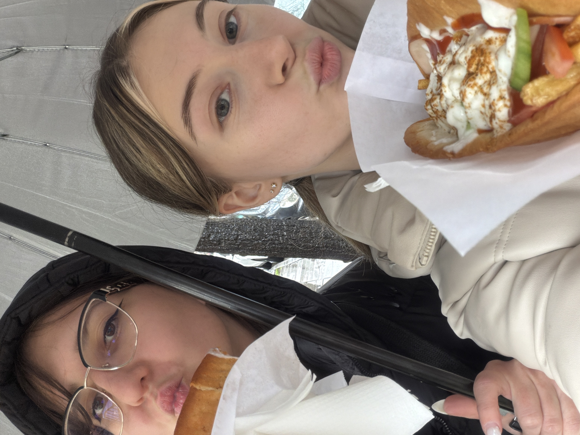Two young women are taking a selfie under an umbrella. The woman at the top has blonde hair with a lighter streak, is wearing a light-colored jacket, and is holding a sandwich with white sauce, ketchup, vegetables, and french fries. The woman at the bottom is wearing glasses and a black hooded jacket, and is also holding a sandwich in white paper. The background includes an umbrella and a glimpse of buildings. The location is Plovdiv, Bulgaria.