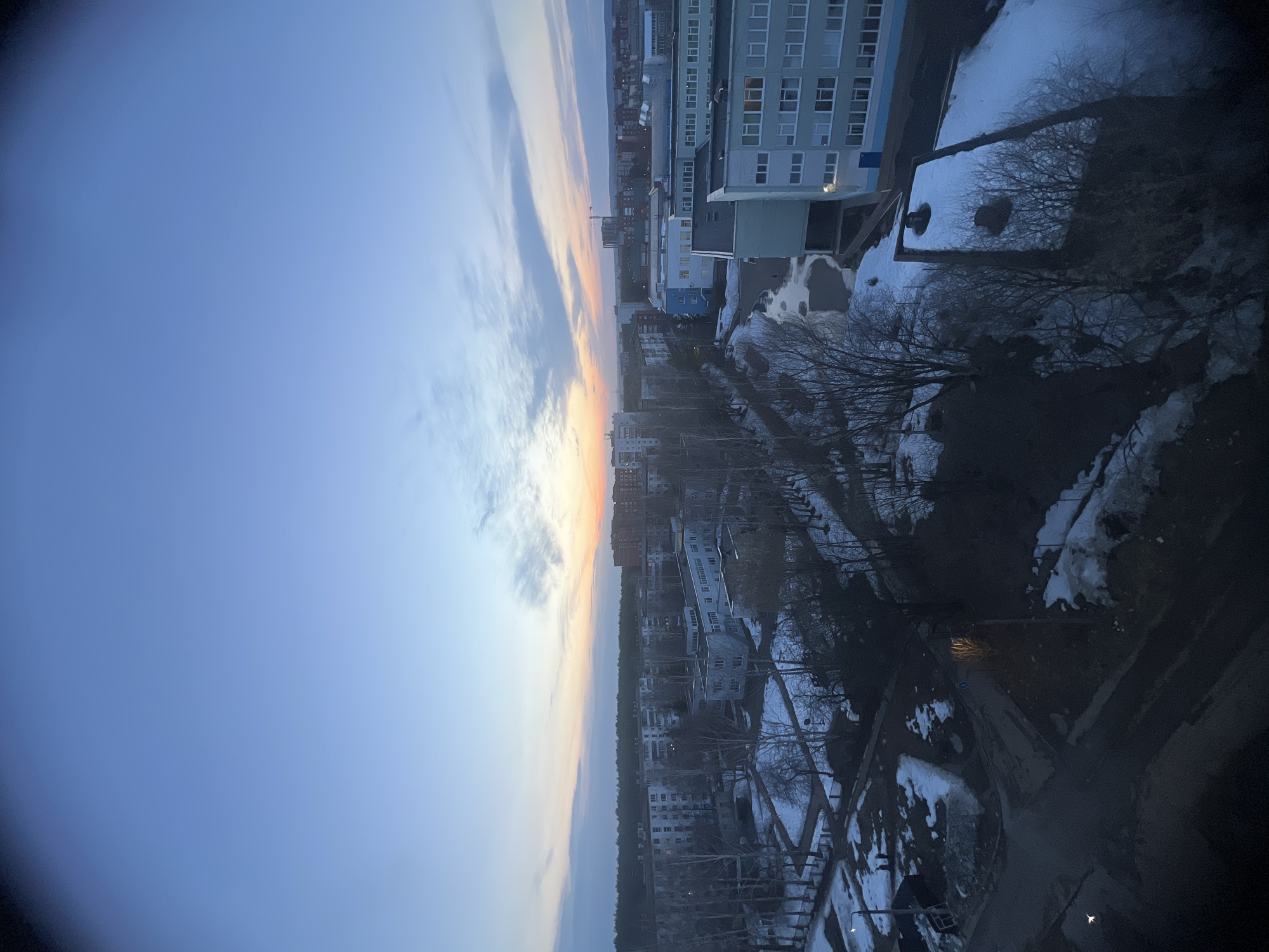 The image shows an elevated, westward view of a cityscape at twilight, most likely in the city of Chaikovsky, Russia, based on the style of the buildings. The sky exhibits a gradient from pale blue to orange near the horizon, with sparse clouds. Below the sky is the city. Multi-story buildings are arranged along a street, some with snow visible on the ground around them. There is also a patch of trees. Patches of snow are present in shaded areas near the foreground. A street is visible with a street lamp. A corner of the building where the photo was shot can be seen in the lower-right portion of the image. No people are readily visible in the scene.