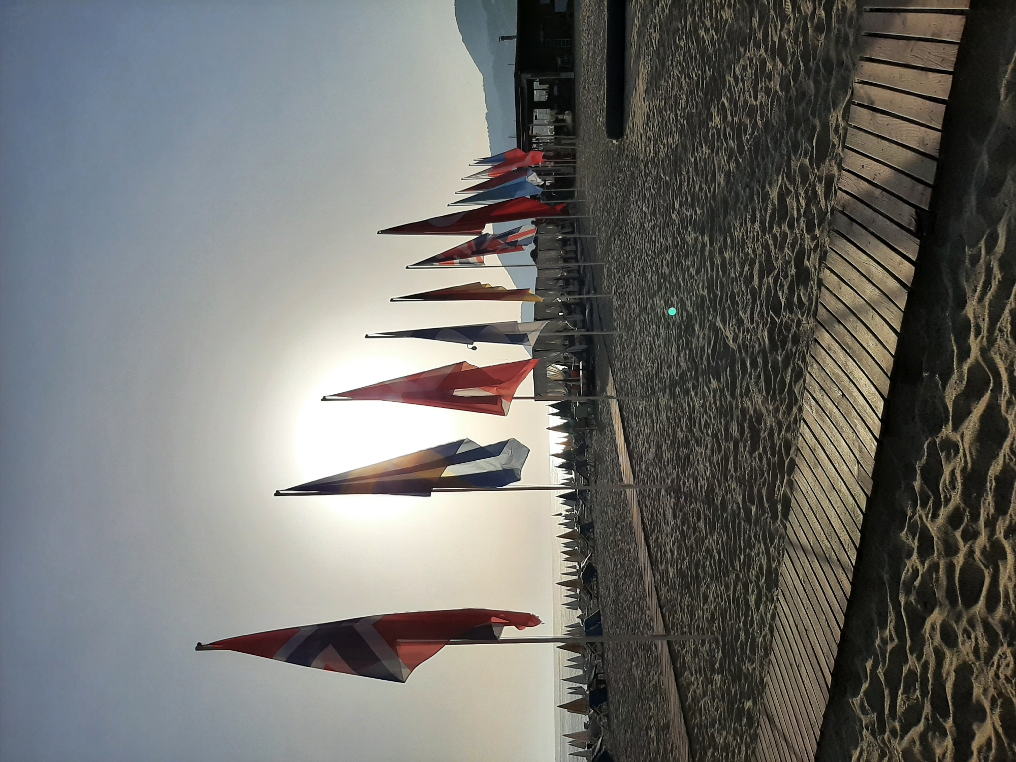 A beach in Alanya, Turkey, features a line of flags stretching into the distance along the sand. The flags are various colors and designs, including some with red and white stripes and others with blue and white patterns. The sun is positioned behind the flags, creating a bright glare in the sky. A wooden walkway runs parallel to the beach. In the background, a building is visible with a mountain looming behind it. Beach umbrellas and lounge chairs can be seen further down the beach. The sand is light brown and textured.