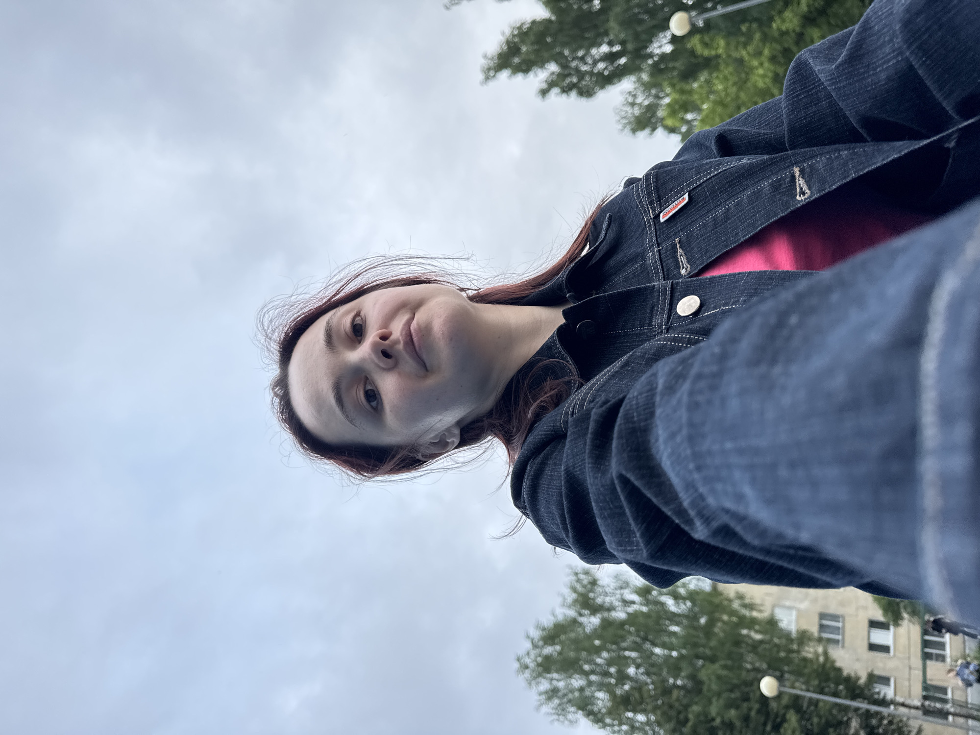 A low-angle selfie captures a young woman with reddish-brown hair looking directly forward. She wears a dark blue denim jacket with white topstitching over a pink top; a red and white fabric tag is attached to the right lapel of the jacket. Her right arm, holding the camera, is visible in the lower foreground. The environment is an urban setting in Izhevsk, Russia, under an overcast sky. The background features blurred green trees on both sides, a multi-story, light-colored residential building with multiple windows, and two streetlights with spherical luminaires. Two indistinct human figures are present near the building in the distance.