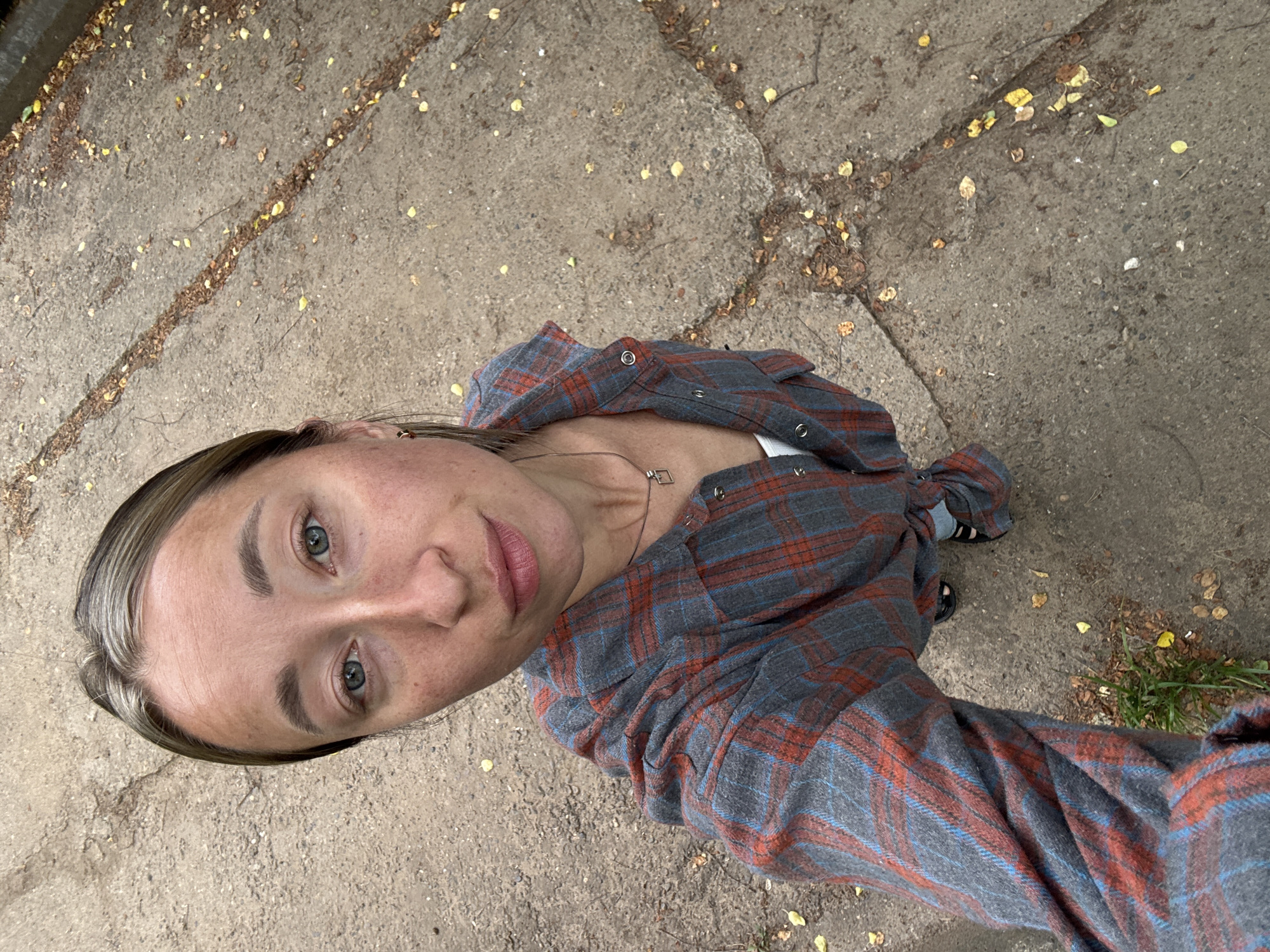 A woman is pictured in a high-angle selfie on a concrete surface. The concrete is segmented into slabs and shows signs of wear and scattered organic debris, likely fallen leaves. The woman's face is prominent, with light eyes, fair skin, and light brown hair. She wears a gray and orange plaid shirt. A silver necklace is visible. Her feet, clad in dark footwear, are at the edge of the frame. The location is Chaikovsky, Russia. No specific action or interaction is apparent.