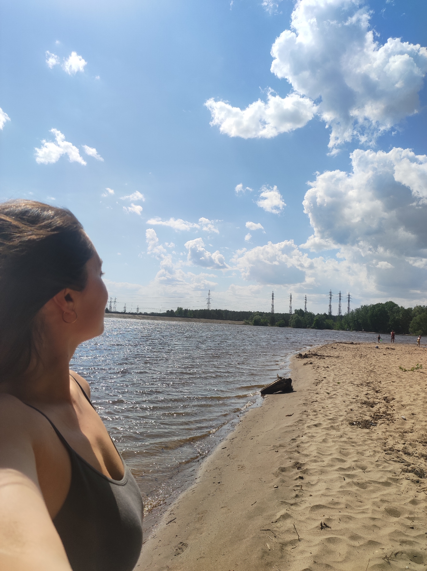 A woman stands on a sandy beach beside a body of water, likely a lake or wide river, with a view of the opposite shore. The sky is blue with scattered puffy white clouds. Several power line towers are visible in the distance along the shoreline. Three people are further down the beach, close to the treeline of the opposite bank. A piece of driftwood rests on the sand near the water's edge. The location is identified as Noviy, Russia. The woman in the foreground is wearing a dark-colored tank top.