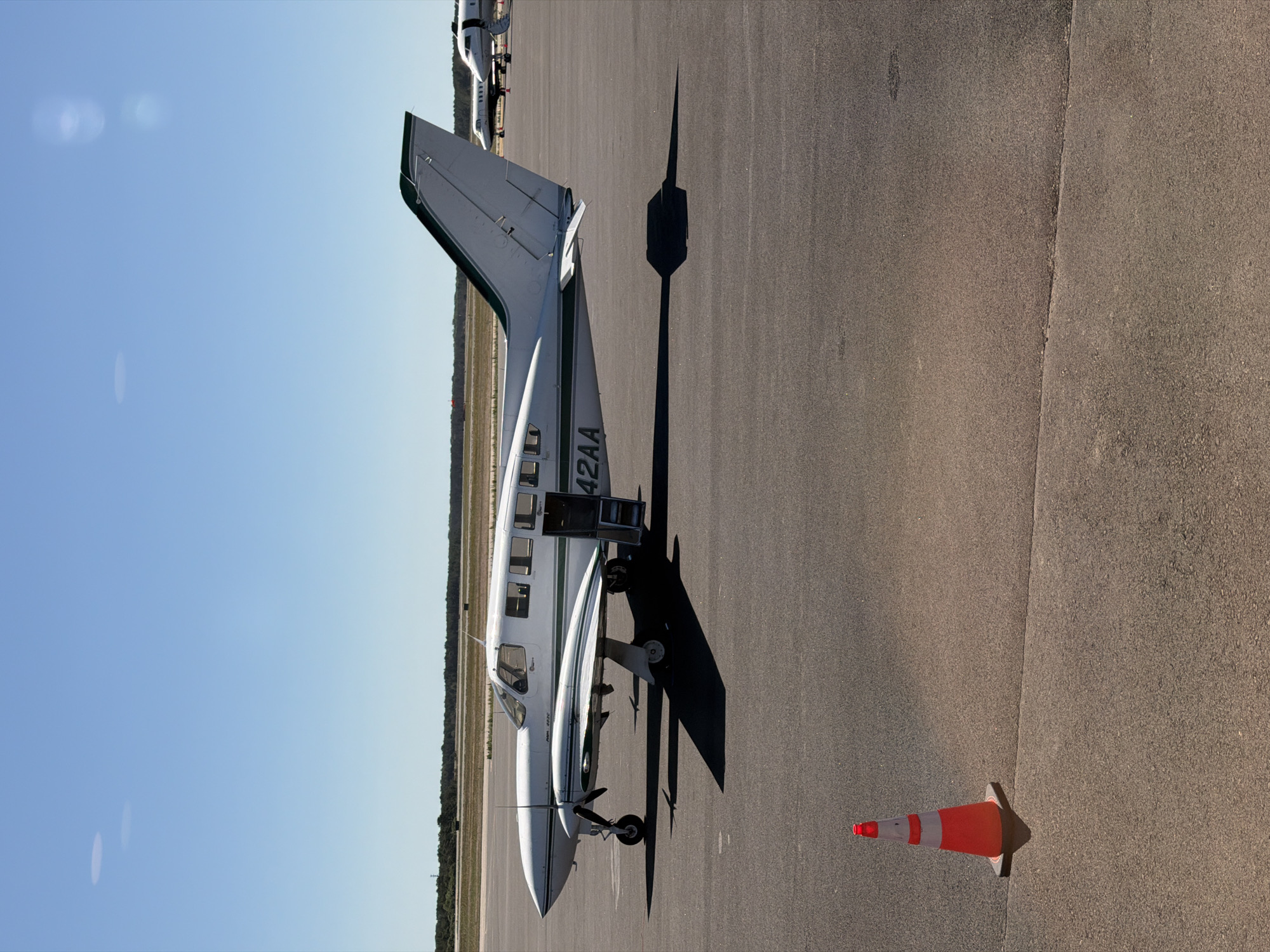 The image depicts a white and green twin-engine propeller aircraft parked on an asphalt tarmac under a clear blue sky. The aircraft's registration number, "42AA", is visible on the tail. The passenger door is open, with steps extended. A red and white traffic cone is positioned in the foreground to the left of the aircraft. In the background, other aircraft are visible, and a line of trees and vegetation forms the horizon. The scene appears to be an airport, likely in a suburban or rural area.