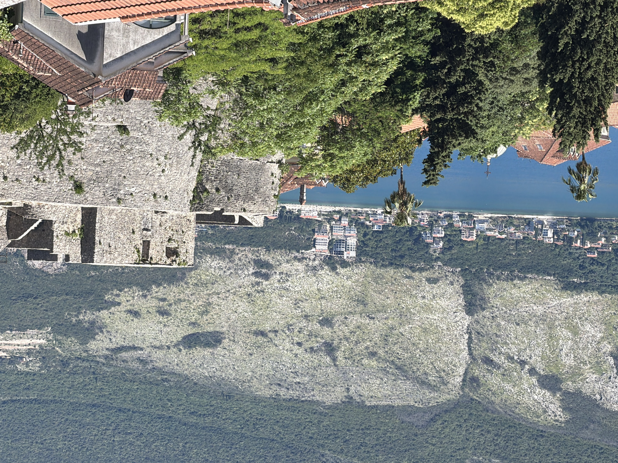 The image shows an elevated view of Herceg Novi, Montenegro. The camera is angled downwards, capturing a landscape of buildings, trees, mountains, and the coastline. In the foreground, there is a stone structure with visible brickwork. Adjacent to this structure are buildings with tiled roofs, interspersed with green trees. In the distance, there is a coastal area with houses and buildings along the shoreline. The coastline is bordered by blue water, and beyond the coastal area, a mountainous terrain covered with vegetation extends into the distance. The sky is not visible in the image.