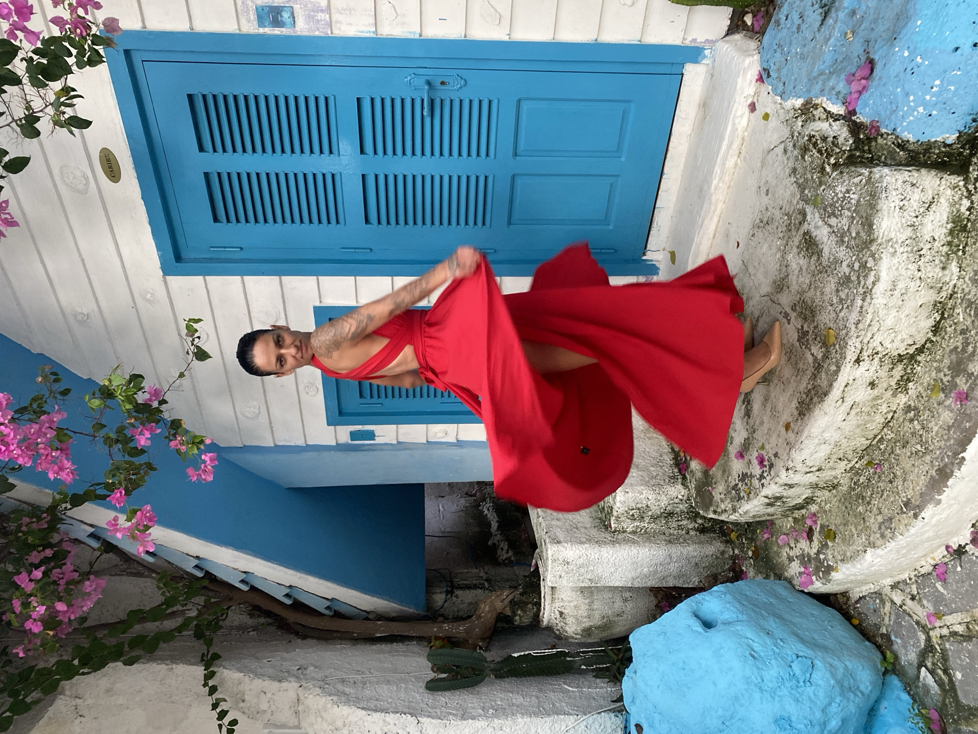 A woman in a red dress stands in an outdoor setting in Phu Thuy Ward, Vietnam. She is facing the viewer and appears to be twirling, causing the dress to flare out around her. The location is characterized by white buildings with blue trim. There is a closed blue shuttered window in the background. Flowering bougainvillea vines with pink blossoms are visible on the left side of the frame. The ground is a mix of concrete and stone, and there are some small, brightly painted blue rock-like features. The overall composition suggests a street scene in a Mediterranean-inspired style.