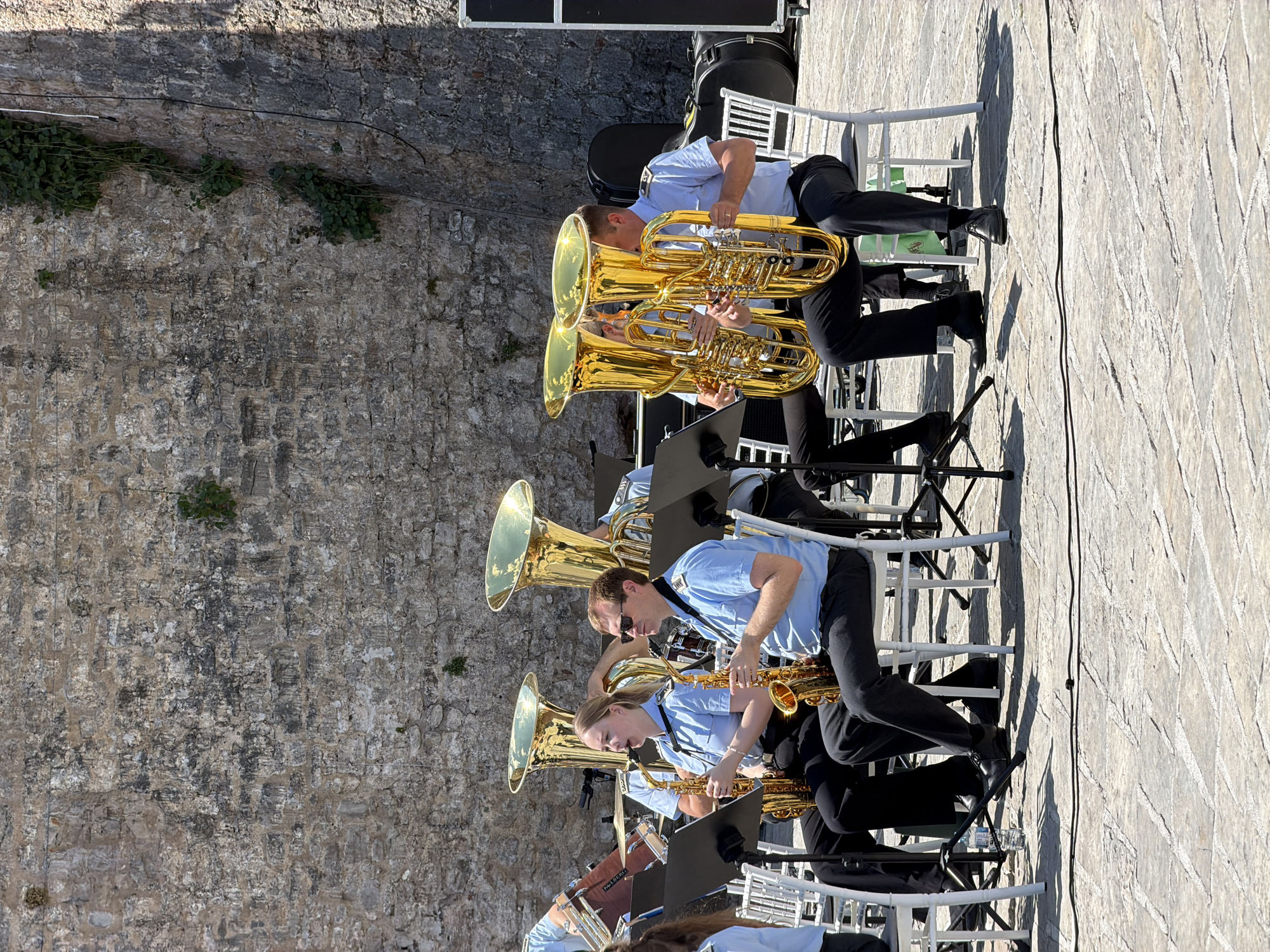 A group of musicians are performing outdoors in what appears to be Boreti, Montenegro. Four musicians are visible, seated on white chairs with music stands in front of them. Two are playing tubas, one is playing a saxophone, and a drummer is partially visible. The musicians are wearing matching light blue short-sleeved shirts and dark pants. The background consists of a stone wall and a paved area. Stage equipment is partially visible behind the musicians.