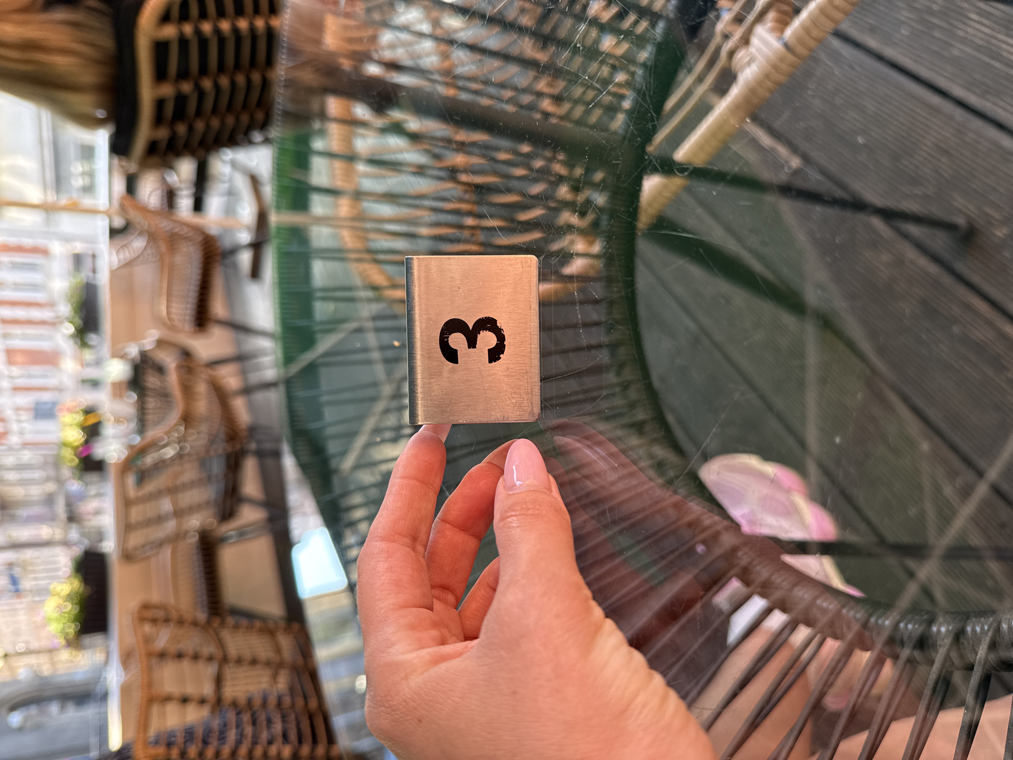 A close-up, high-angle shot captures a left hand holding a metallic table number "3" on a glass-topped outdoor table. The hand has light-toned skin and neatly manicured nails with a light pink polish. The table number is a brushed metal, rectangular block with a black, slightly distressed "3" printed on one side. The glass table surface shows numerous scratches and reflects light, revealing the underside structure of a wicker chair and a wooden deck floor below.
In the mid-ground, blurred details of an outdoor seating area are visible. Several light brown, woven wicker chairs with metal frames are arranged around similar glass tables. Beyond the seating, there are dark planters holding green foliage and bright, colorful flowers. The background features more heavily blurred architectural elements, suggesting buildings with windows, typical of an urban street or square. A person with blonde hair is partially visible in the far right background, seated in one of the wicker chairs. The lighting suggests a bright, possibly sunny, daytime environment. The scene depicts a casual outdoor dining or cafe setting in the city of Unknown, Unknown.
