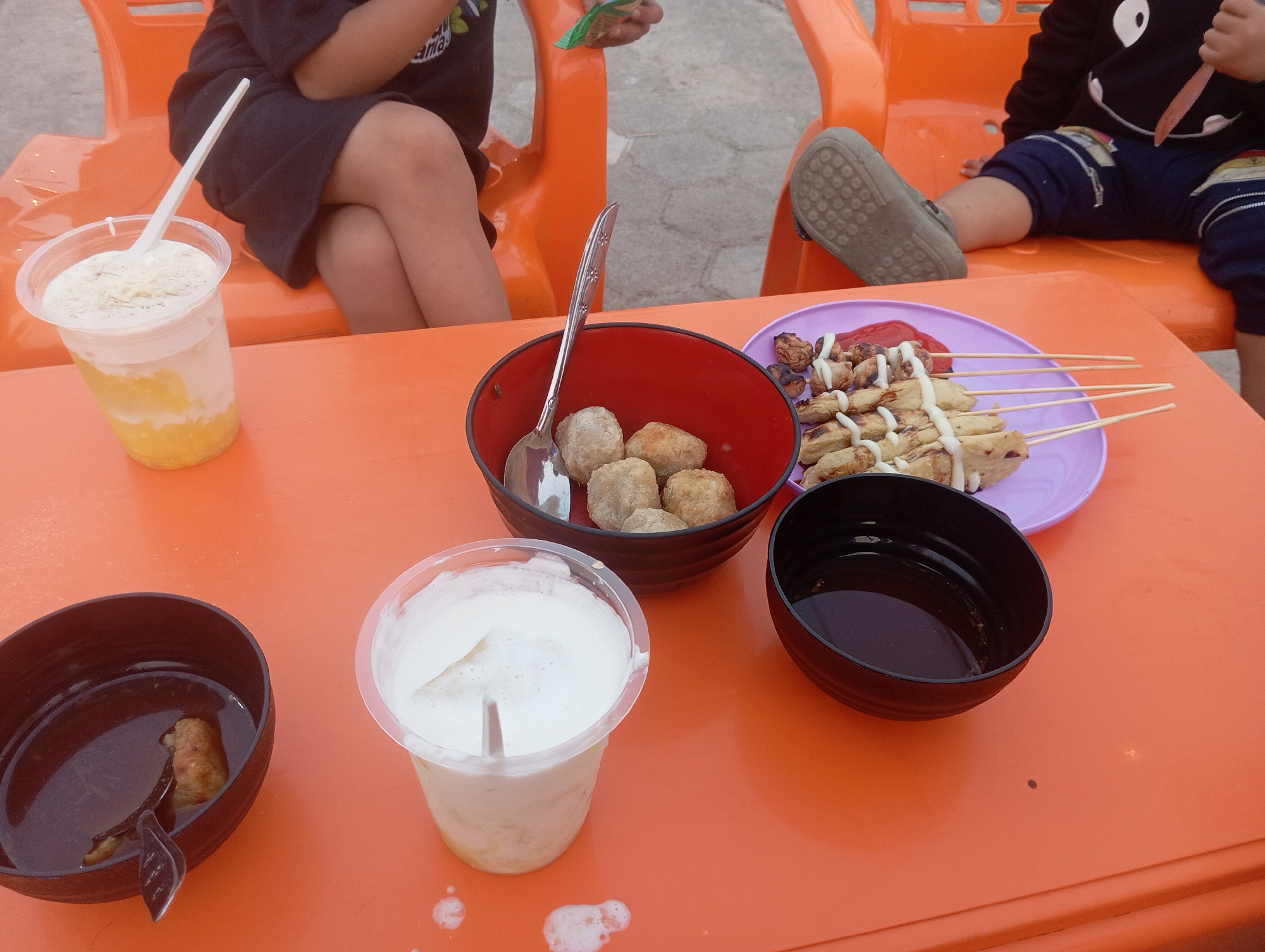 In Way Dente, Indonesia, an orange plastic table holds a variety of food and drinks. In the foreground, two plastic cups of beverage, one with a white frothy top and the other with a yellow and white layered liquid, are partially visible. To the left, a black bowl contains a small amount of liquid and a piece of food, with a spoon inside. To the right, a red bowl is filled with several round, light-brown food items and has a silver spoon resting on the rim. A purple plate holds skewers of cooked food topped with white sauce and red sauce. In the background, two orange plastic chairs are occupied by two individuals. One person, wearing a dark shirt and skirt, sits with their legs crossed. The other person, in a black hooded sweatshirt with a white design on the front, has their legs extended. One of the individuals is holding a small green packet.