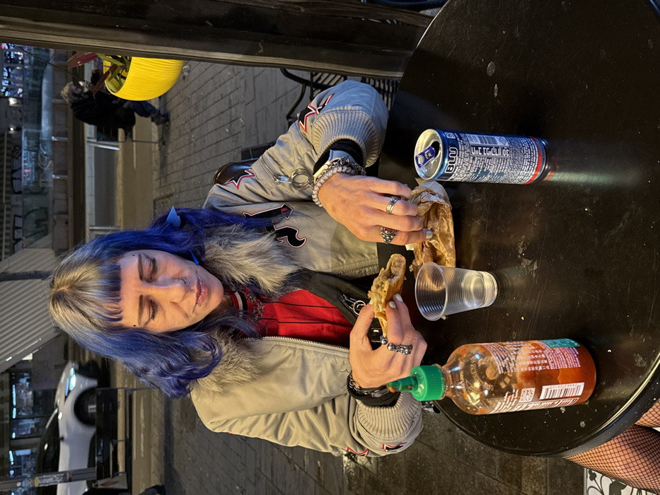 A person with blue hair and multiple piercings sits at a black table outdoors. They are wearing a gray jacket with a fur-lined collar and a red shirt. On the table, there's an open can of a blue energy drink, a partially eaten taco in paper wrapping, a clear plastic cup, and a bottle of Sriracha hot sauce. The person is adding Sriracha to the taco. A blurred street scene is visible in the background.