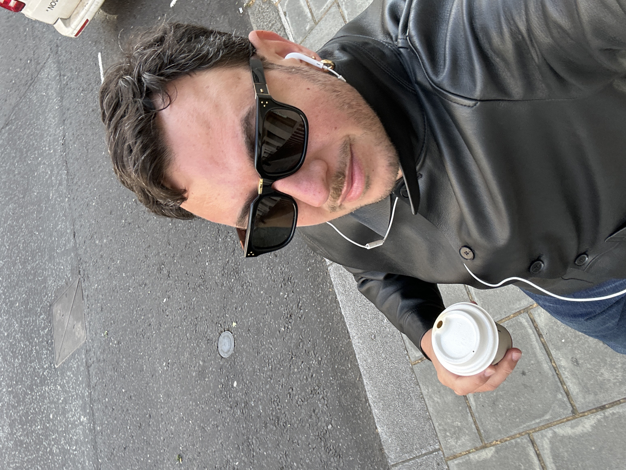 The image is a high-angle selfie of a man, rotated counter-clockwise, portraying a casual urban scene, likely in London, United Kingdom. The man is looking down towards the camera, wearing black sunglasses with gold accents on the frames and white wired earphones. He has dark, wavy hair with some visible grey strands and light facial stubble. He is dressed in a black leather jacket over a dark shirt, with a portion of blue jeans visible. In his right hand, he holds a white disposable coffee cup with a lid.
The setting is an outdoor street or sidewalk. The ground beneath him is a mix of dark grey asphalt speckled with lighter aggregate and lighter grey rectangular paving stones. Embedded in the asphalt are small, circular and diamond-shaped metal plates. In the top left corner, the rear section of a white vehicle, possibly a van or car, is partially visible, parked on the asphalt. The word "NOV" is visible on the side of this vehicle. The lighting indicates it is daytime, and the overall atmosphere is casual, suggesting the man is either walking or paused during a daily activity like getting coffee.