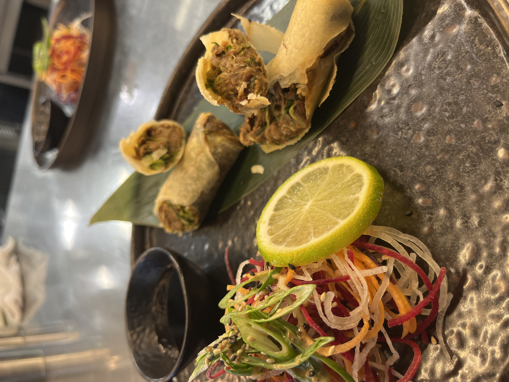 This image captures a close-up, slightly angled view of a plated meal, likely an appetizer, in what appears to be a restaurant setting.
The main subjects are several spring rolls, a vibrant side salad, and a slice of lime, all arranged on a dark, textured plate. Two of the spring rolls are prominent; one is cut in half, revealing a savory filling of shredded, dark meat (possibly duck or pork) mixed with green herbs or vegetables. Its wrapper is golden-brown and looks crispy. Another spring roll, mostly intact, lies beside it on a long, slender green leaf, which serves as a decorative element. A smaller, cut piece of a spring roll is visible to the left.
Adjacent to the spring rolls is a colorful salad made of finely shredded vegetables, including red, orange, and white strands. This salad is garnished with sliced green onions and sprinkled with both black and white sesame seeds, suggesting an Asian-inspired dish. A fresh, bright green lime slice rests on top of the salad, adding a pop of color and indicating a potential citrusy flavor element.
In the lower left corner, a small, dark, cylindrical bowl is visible, likely intended for a dipping sauce. The plate itself is dark grey or charcoal in color, with a rustic, slightly dimpled or textured surface.
The background is out of focus, showing glimpses of another plate with food, suggesting a larger dining environment. The lighting appears to be artificial, typical of an indoor restaurant setting. No visible text is present in the image. While the location is given as Sofia, Bulgaria, there are no specific visual cues within the image to confirm this.
