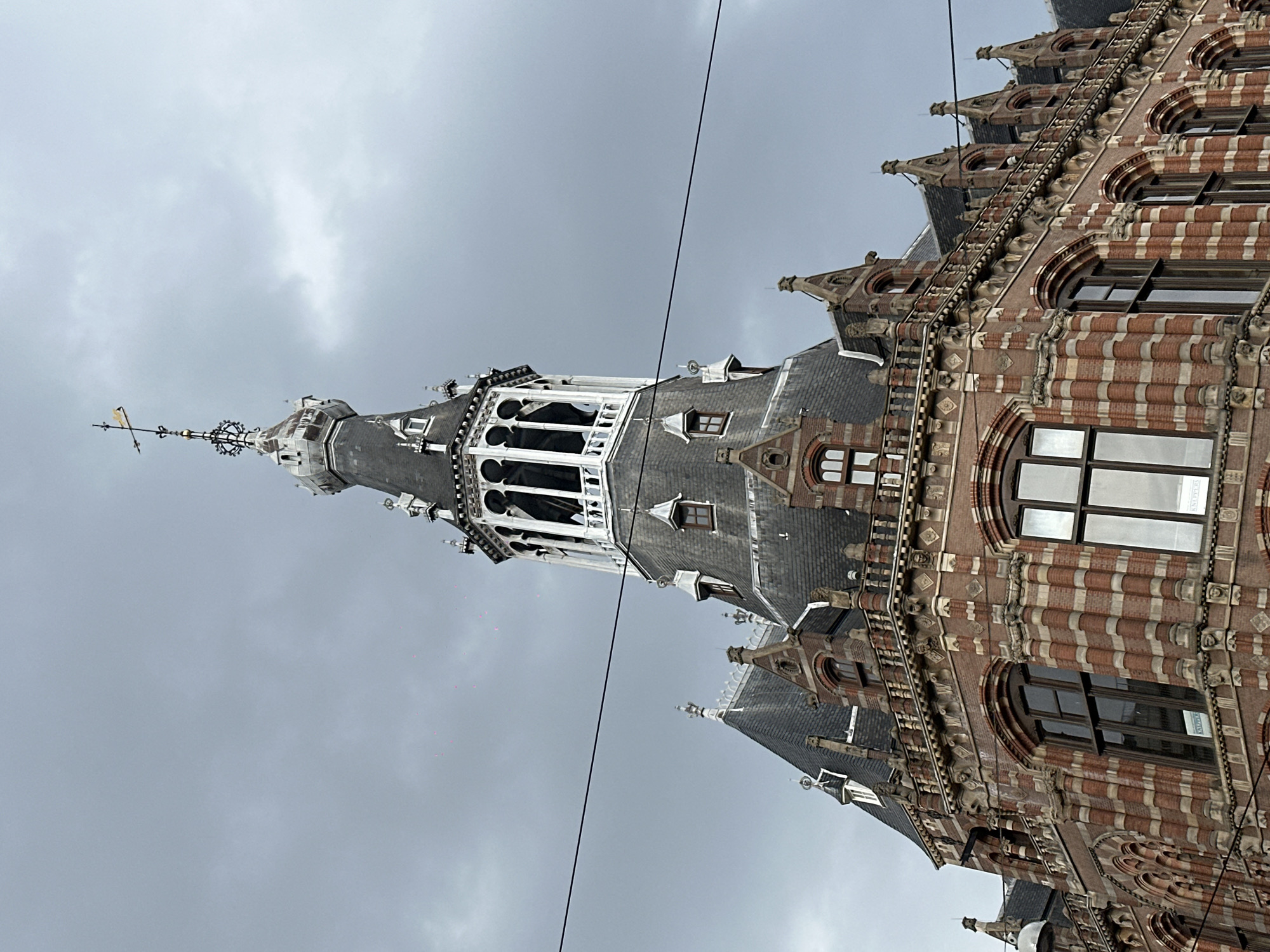 The image captures the upper portion of the Grand Hotel Amrâth Amsterdam, a historic building located at Prins Hendrikkade 109 in Amsterdam, Netherlands. The architecture is characterized by its intricate brickwork in red and light sandstone, with numerous decorative elements, arched windows, and steep, dark grey slate roofs. A prominent tower with a white, open-air lantern section topped by a weather vane dominates the center of the frame. The sky is overcast and grey, suggesting a cloudy day. Two thin black wires run horizontally across the image, obscuring parts of the building's facade. No people are visible in the frame.