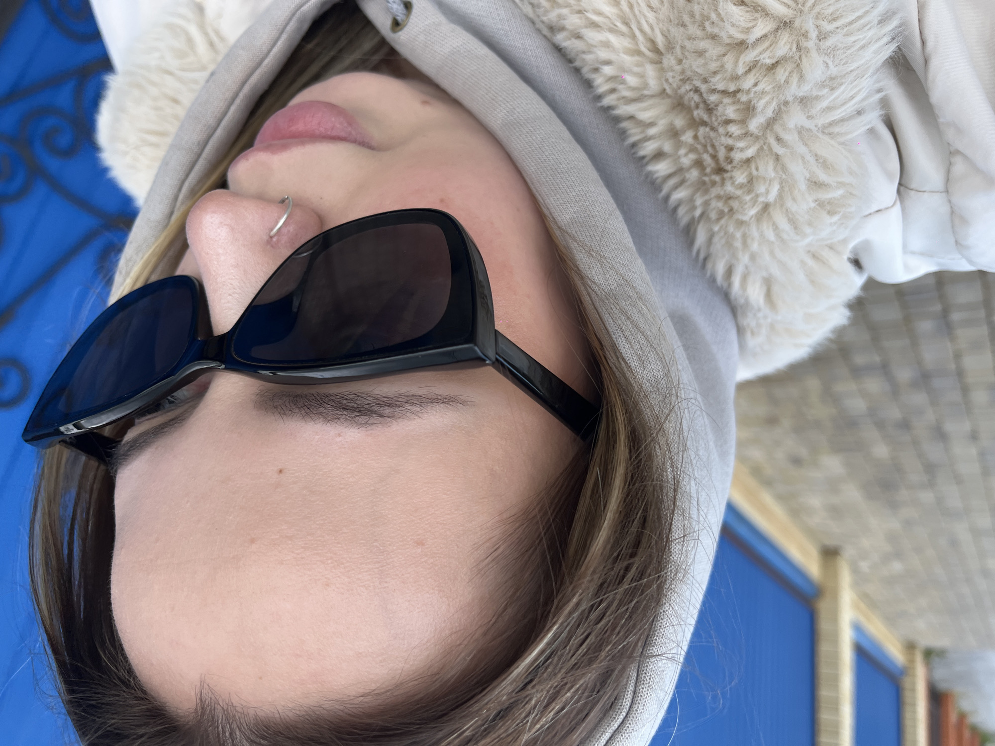 A close-up, rotated portrait shows a person wearing sunglasses and a hooded jacket. The person has a nose ring. The background shows blue and beige structures, consistent with urban architecture. The exact location within Novopokrovskaya, Russia, cannot be determined with only the visual data provided.