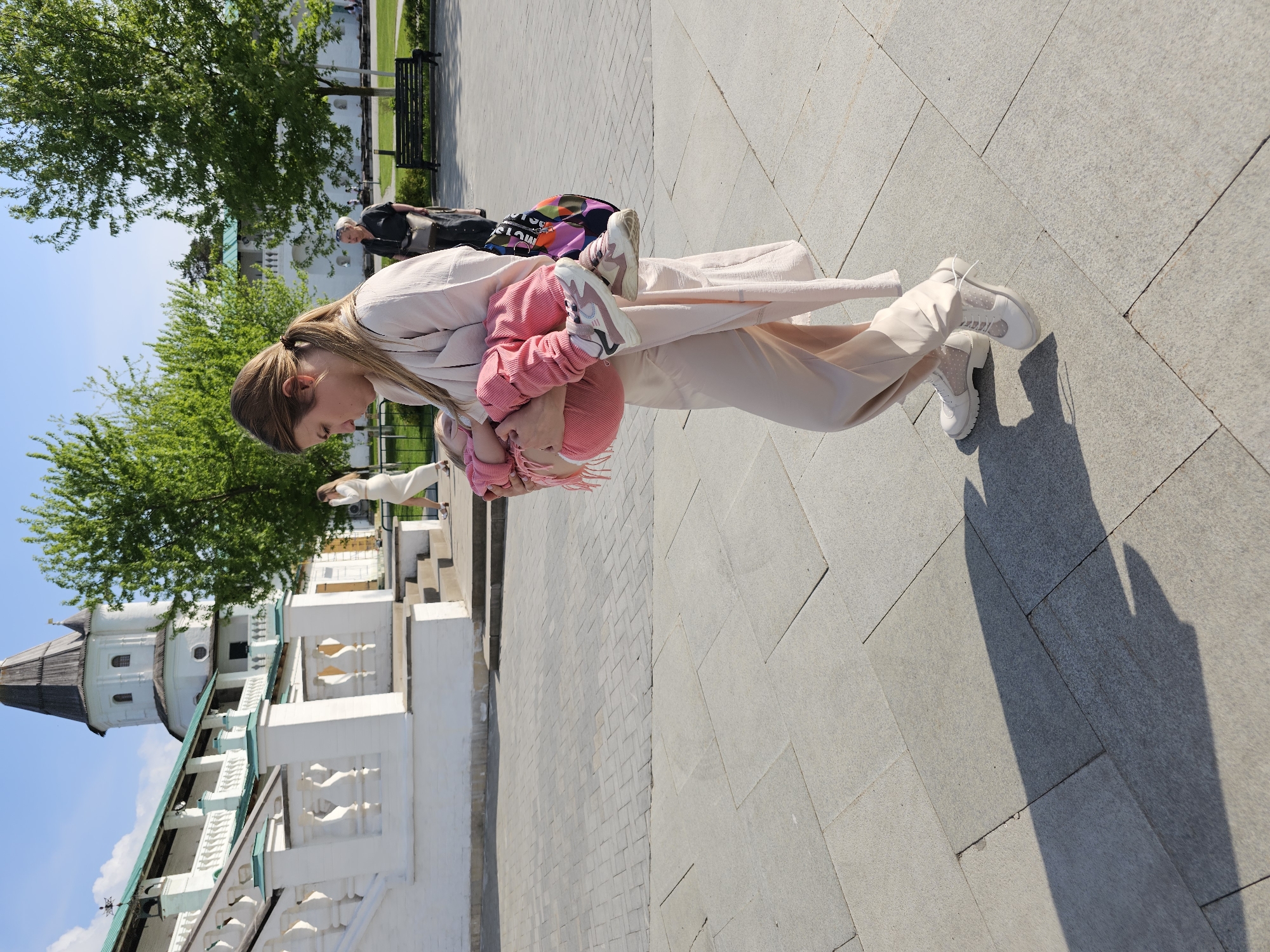 A woman wearing a light tan outfit and white sneakers is holding a toddler, dressed in pink, in her arms outdoors. The woman is bending slightly forward, looking at the child. To the left of the woman, a white building with a green roof and a tower is visible. Lush green trees are in the background. Paved ground stretches across the foreground and background. Another person walks on the upper right.
This scene takes place in Istra, Russia.