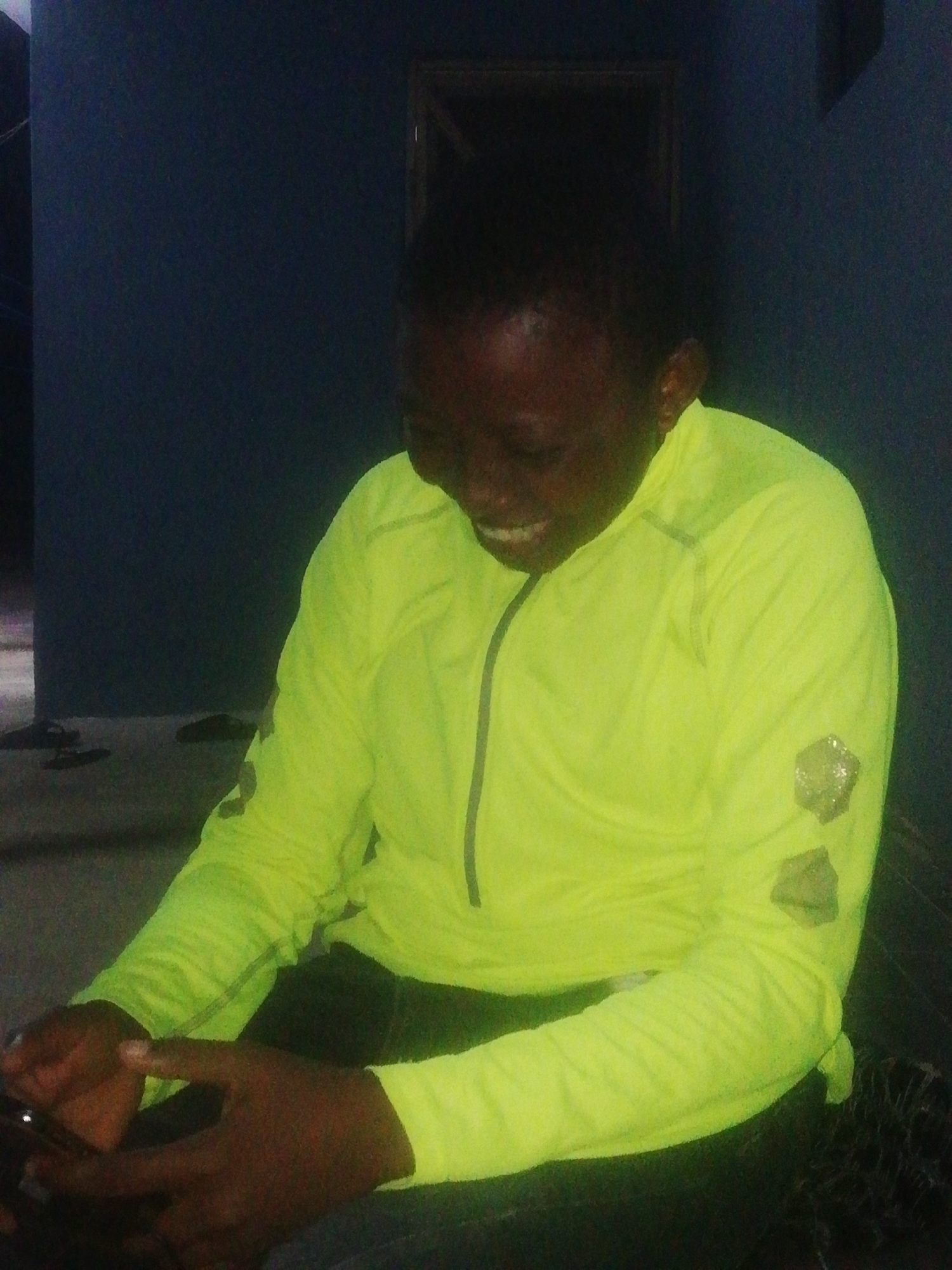 A person with dark skin and short hair is sitting indoors. The person is wearing a bright yellow long-sleeved shirt with a zipper at the collar and dark pants. The person is looking down and smiling. The person is holding a cell phone with both hands. The interior of the location seems to be a room with a dark blue wall, a mirror, and some objects on the floor, potentially sandals or shoes. The location is in Ikorodu, Nigeria.