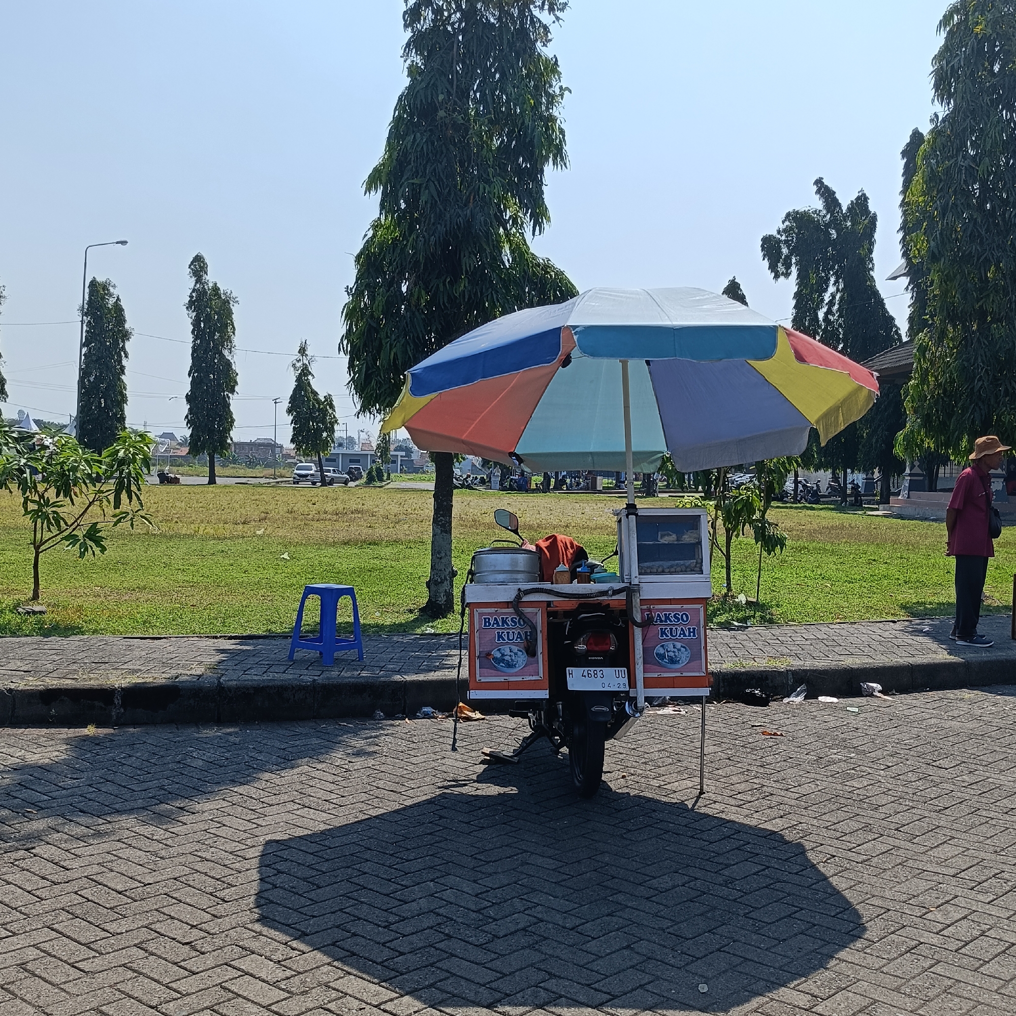 An outdoor scene in Kendal, Indonesia, features a food cart selling "Bakso Kuah" (meatball soup) parked on a paved area. The cart is attached to a motorcycle and is covered by a colorful umbrella. A blue plastic stool is placed to the left of the cart, and a person wearing a straw hat and maroon shirt stands to the right, looking away from the camera. The background consists of a grassy field with several tall trees, a sidewalk, and a few vehicles parked in the distance. The sky is clear and bright, suggesting a sunny day. The scene appears to be in a park or a public space.