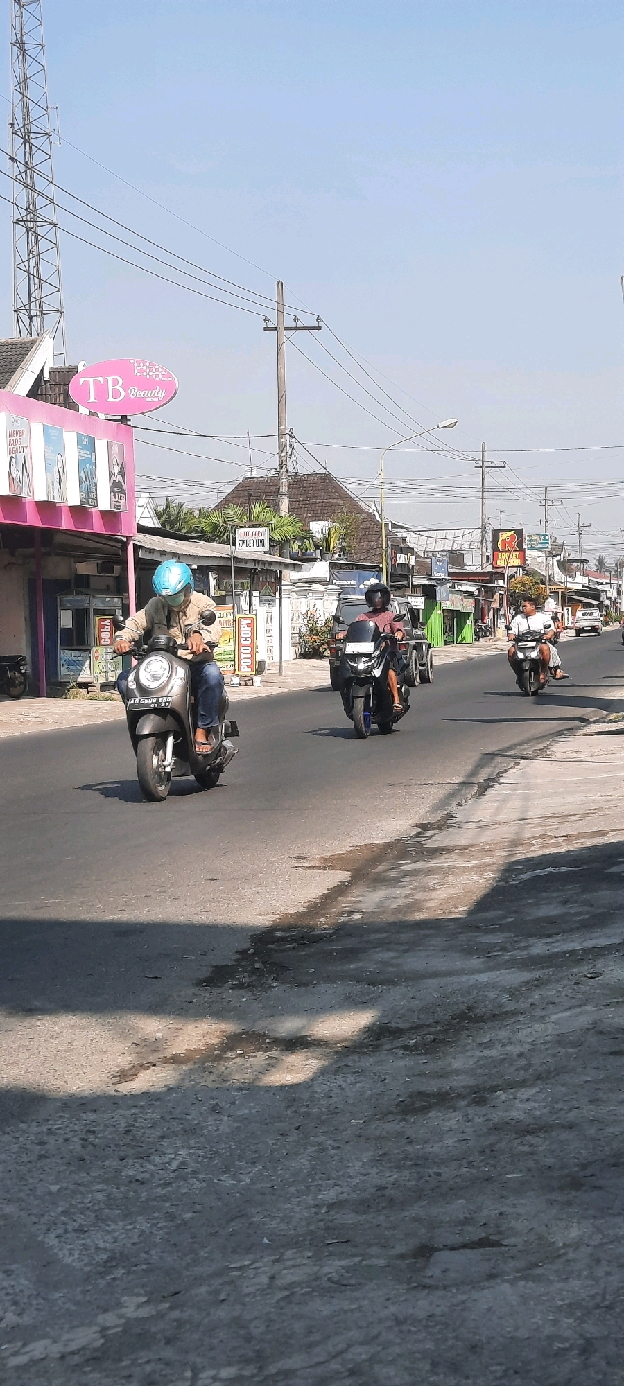 The image shows a street scene in Selopuro, Indonesia, under a clear blue sky. A cell tower is visible on the left side of the frame, alongside a building with a pink facade and a sign that reads "TB Beauty Store." Several people are riding motorcycles on the asphalt road. In the foreground, a person in a light brown jacket and blue helmet is riding a white scooter with license plate "AC 6608 BBD." Behind them, another person on a black scooter is also visible. Further down the road, more motorcycles and a white car are present. The roadside features various buildings, some with awnings and signs, and utility poles with electrical wires. Shadows are cast across the road, indicating sunlight.