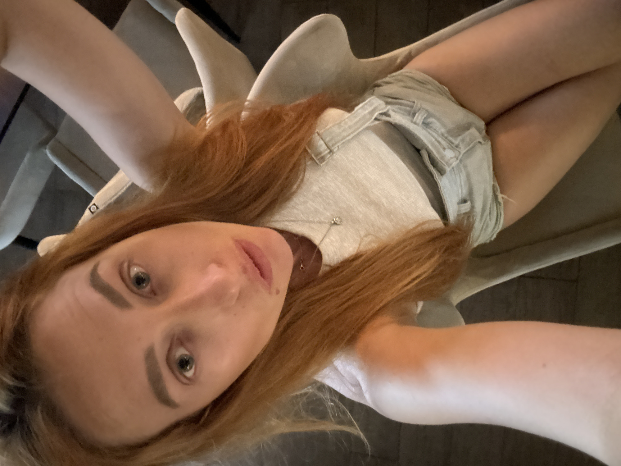 A woman with auburn hair and fair skin is taking a selfie while sitting in a chair. She is wearing a light-colored tank top and denim overalls. The background shows a dimly lit interior, possibly a restaurant or lounge, with other chairs visible. The woman's face is close to the camera, and she is looking directly at it.