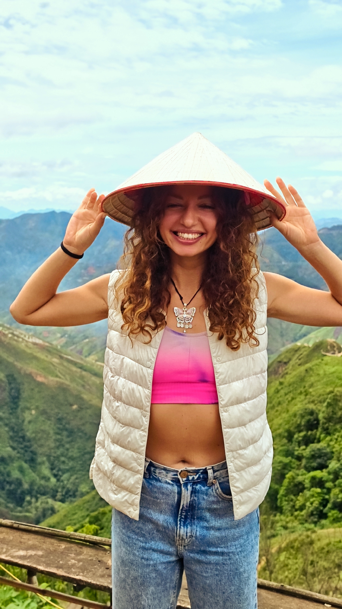 A young woman stands outdoors wearing a conical hat and a white puffer vest over a pink crop top and blue jeans. She is smiling with her hands raised to the sides of her hat. The background shows a lush, mountainous landscape with some clouds in the sky. The scene appears to be a viewpoint overlooking a valley. This location is likely in or around Ta Xua, Vietnam, a region known for its natural beauty and mountainous terrain.