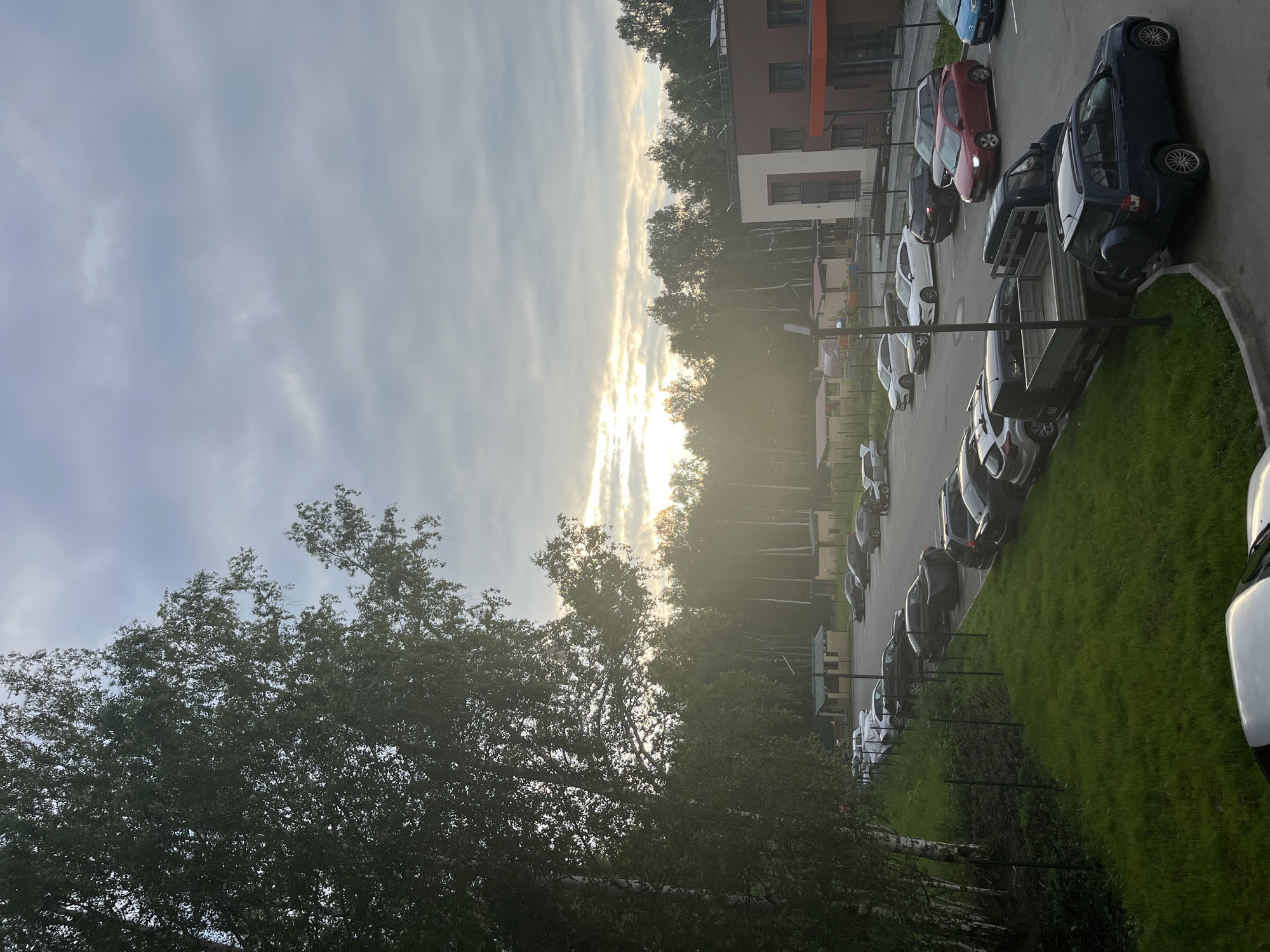 The image depicts a residential area in Prigorodny, Russia, under an overcast sky with patches of sunlight breaking through. In the foreground, a lush green lawn slopes down towards a paved parking lot filled with various cars. A dark blue SUV is parked in the lower right of the frame, facing left. Adjacent to it, a flatbed truck is parked, facing left, with its cargo bed empty. Behind the truck, several other vehicles are parked, including a red sedan, a blue hatchback, a black SUV, and a white hatchback.
To the left of the parking lot, a line of silver birch trees with white bark stands tall, their leaves creating a dense canopy. A wire fence separates the grassy area from a dirt path running alongside the trees.
In the middle ground, a two-story building with brown and white facades is visible, featuring several windows. To the left of this building, smaller structures resembling sheds or garages are present. A line of trees forms a dense backdrop to the scene, with the sun casting a warm glow through them. The overall impression is of a quiet, semi-urban environment with a touch of nature. There are no people visible in the image.