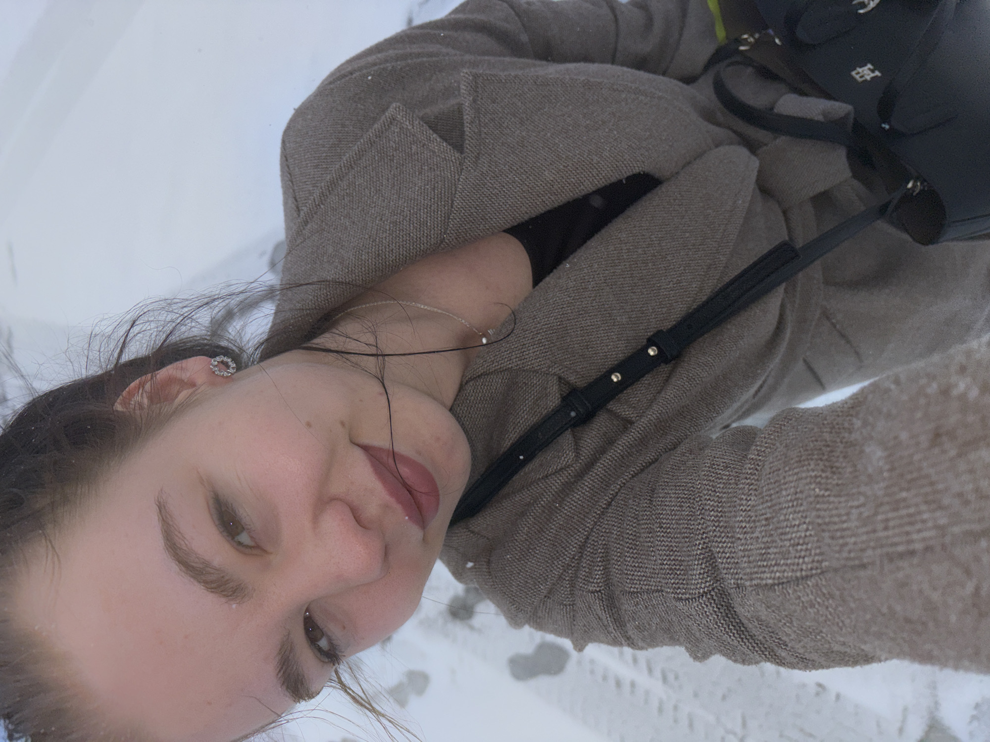 A young woman is pictured in a close-up selfie while outdoors. She is wearing a brown tweed jacket and a black purse is visible on her shoulder. The background is blurry but seems to be a snowy landscape. Considering the metadata is supposed to indicate the location as Perm, Russia, the picture appears to be taken there, likely in a snowy urban environment. The woman has fair skin, light brown hair and is wearing an earring. She is smiling slightly.
