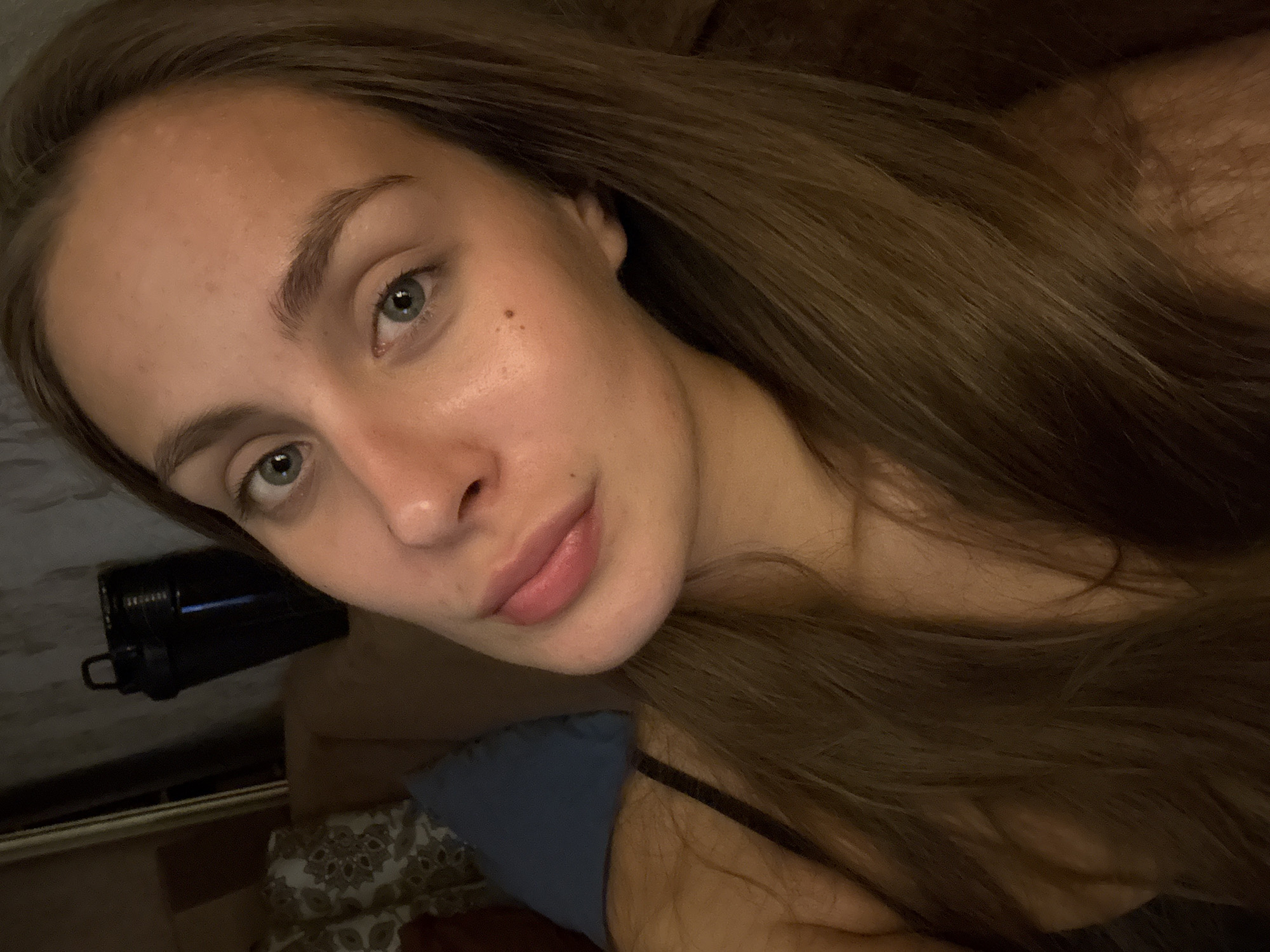 A young woman with long brown hair and blue eyes is taking a selfie. She is wearing a black strap, possibly from a camisole or bra. Her face is smooth with a few freckles visible. In the background, a dark object with a handle is visible, possibly a water bottle or shaker. Behind that, a patterned cushion and a hint of a sofa can be seen, suggesting an indoor setting. The overall lighting is dim and warm, creating a cozy atmosphere. The location is described as being in the city of Шолохово, Russia, but this information is not visually evident from the image itself.