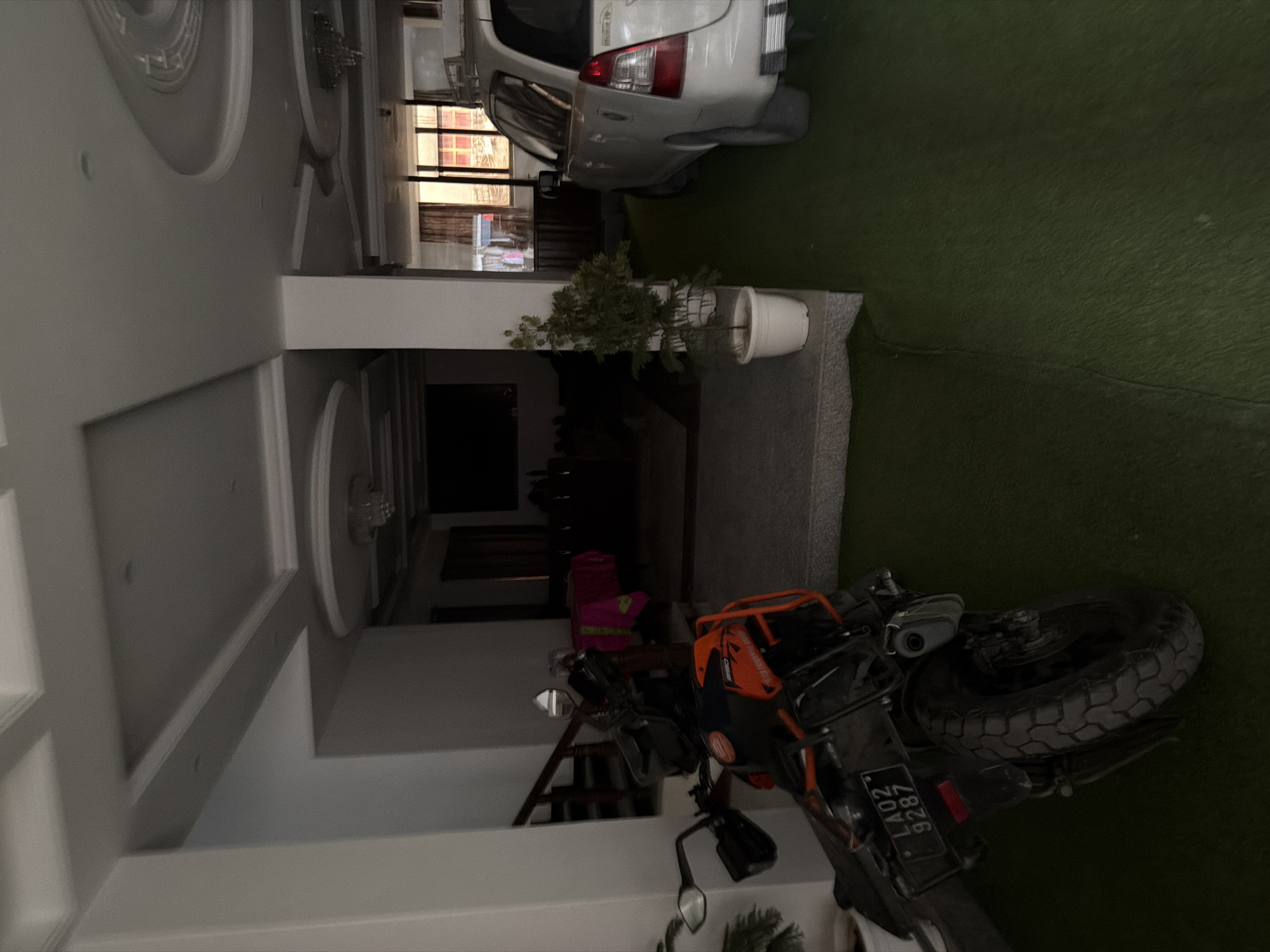 A motorcycle is parked on artificial turf in what appears to be a covered outdoor area or a very large garage. The motorcycle is orange and black and has a license plate that reads "LA02 9287". To the right of the motorcycle is a white SUV, parked and facing away from the camera. Behind the vehicles, there is an indoor area visible with windows, curtains, and some furniture. The ceiling has decorative moldings. The overall impression is of a private residence or establishment with parking. The image is taken in Leh, India.