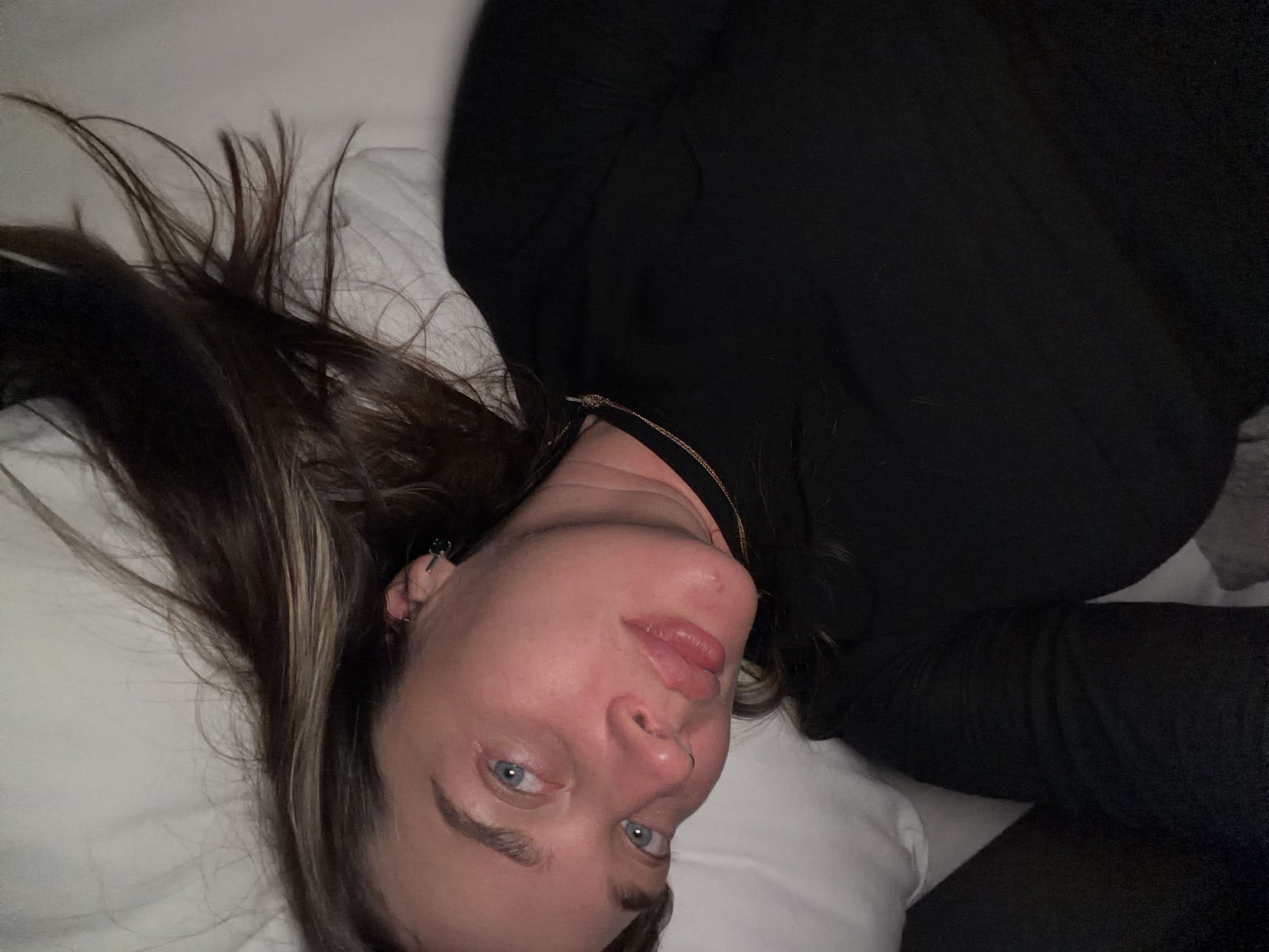 A close-up, overhead, indoor shot shows a fair-skinned woman with medium-brown hair, lying down on a white pillow in Solov'evo, Russia. The woman is wearing a black long-sleeved shirt and a delicate gold necklace. Her blue eyes are open, and her lips are slightly parted. The lighting appears to be artificial and dim, casting a warm glow on her skin. The background consists of white pillows and dark fabric.