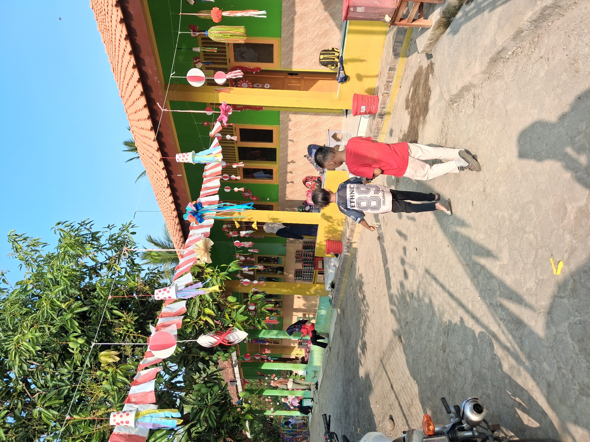 The image depicts an outdoor scene in front of a colorful building, likely a school or community center, in Bantarkawung, Indonesia. The building is painted green with yellow trim and has a terracotta tile roof. Decorations, including strings of red, white, and blue paper ornaments and flags, are hanging from trees and the building's overhang. Two young boys, one in a dark t-shirt with "ETHNIC 89" on it and the other in a red t-shirt, are walking away from the camera towards the building. Several other individuals, including women in headscarves, are visible in the background, some sitting on a low wall and others standing. In the foreground, a portion of a motorcycle is visible on the left. The ground is a dirt path. The overall atmosphere suggests a festive or celebratory occasion.