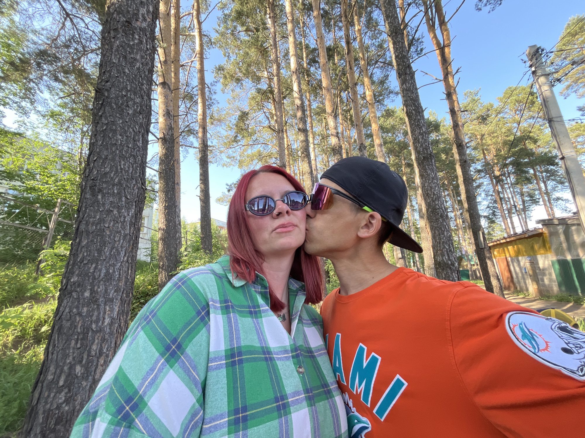 A man and a woman pose for a selfie outdoors in Chaikovsky, Russia. The woman is on the left, wearing a green and white plaid shirt and dark sunglasses. Her hair is reddish-brown. The man is on the right and is kissing the woman's cheek. He wears a black baseball cap, orange long-sleeved shirt with "MIAMI" in teal lettering, sunglasses, and has a Miami Dolphins patch on his left sleeve. Both are wearing sunglasses. They are positioned against a backdrop of tall pine trees and blue sky. There is also greenery to the left, and structures in the background. A utility pole is on the right.