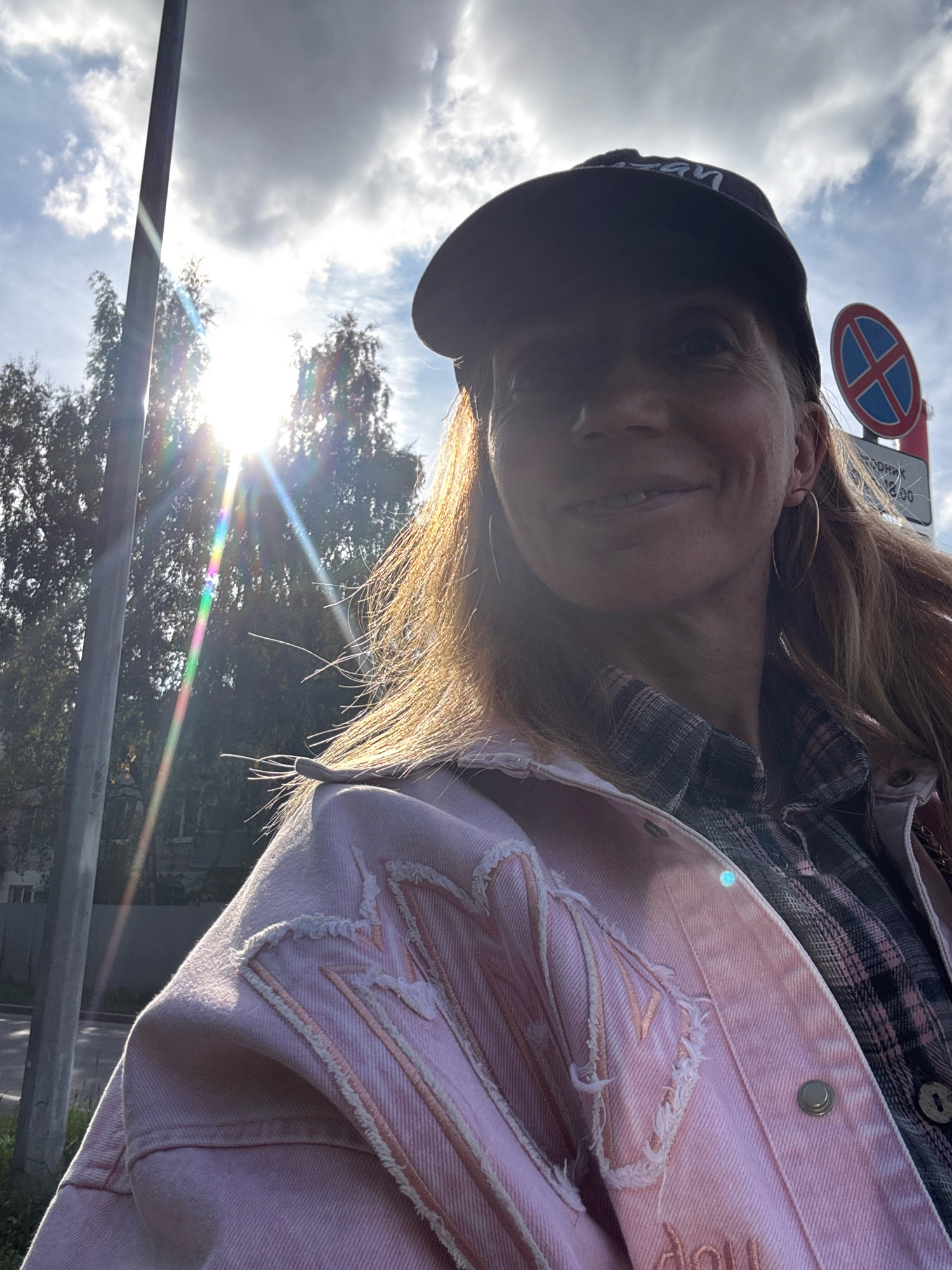 A woman wearing a pink denim jacket over a plaid shirt and a baseball cap is taking a selfie outdoors. Sunlight flares from the left side of the image, creating lens flares and illuminating her hair. A lamppost is visible to the left, and trees line the background. A "No Parking" sign is in the upper right corner, with Russian text below it. The scene appears to be on a street in Ladozhskoe Ozero, Russia.