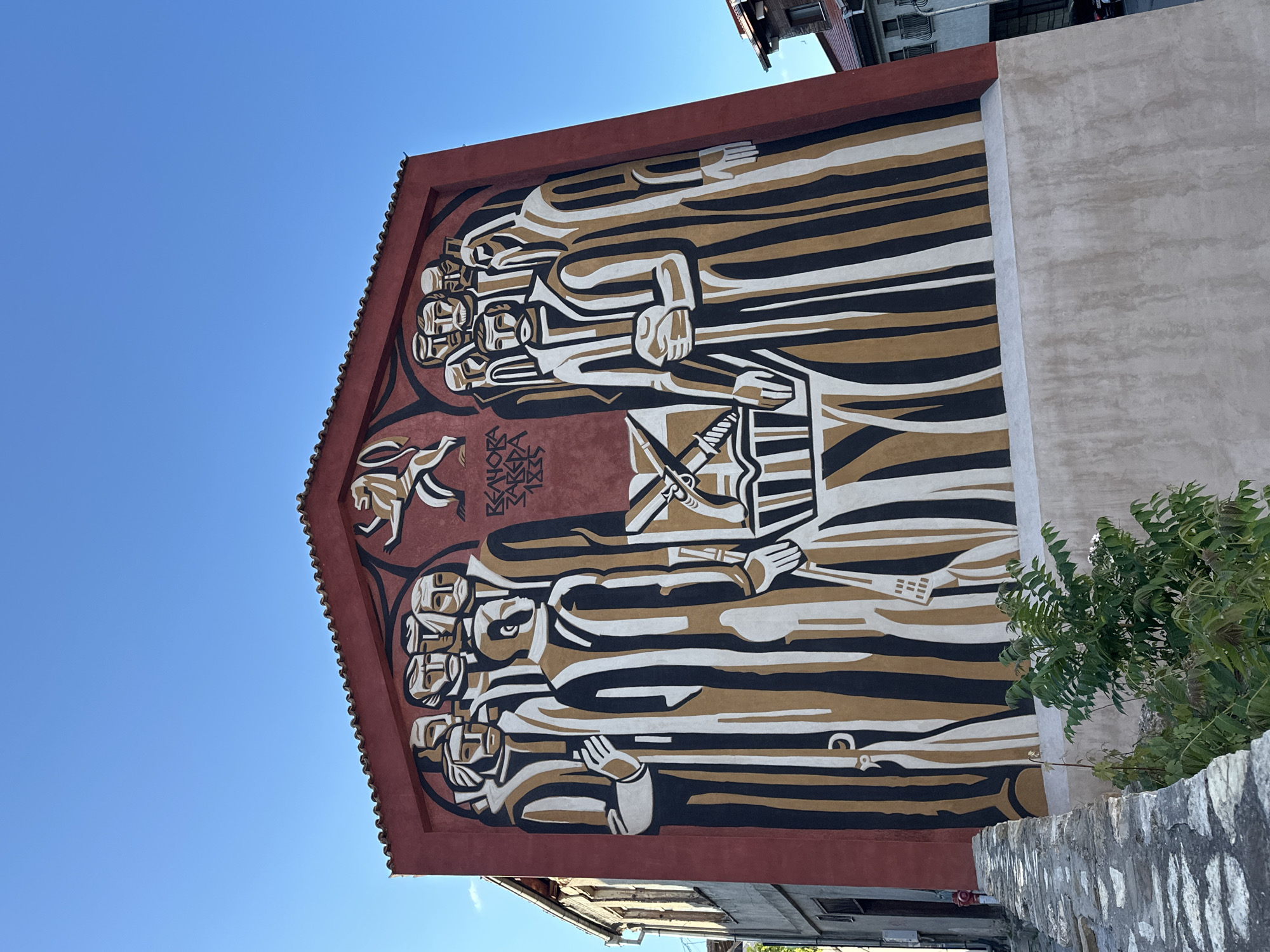 The image showcases a large, stylized mural on the side of a building in Veliko Tarnovo, Bulgaria. The mural is primarily red, gold, black, and white. The composition features rows of stylized figures, depicted in a linear, abstract manner, with an emphasis on their elongated forms and interweaving patterns. Near the top of the mural, a stylized running animal and Cyrillic text are visible. Below the figures is an image of a sword. The building has a red tiled roof. A corner of the building and some trees are in the right foreground. Blue sky is visible above the building.
