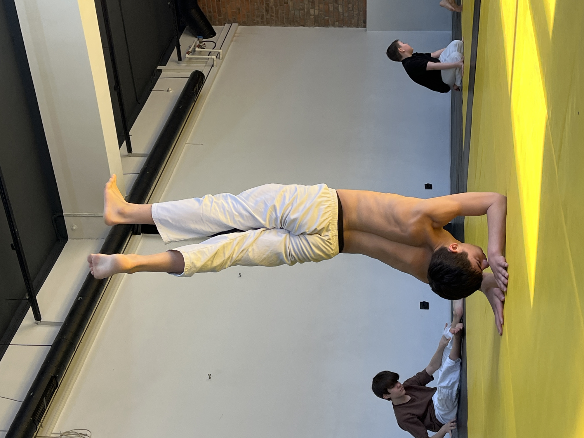 This image captures an indoor training session in what appears to be a gymnasium or martial arts studio in Krasnoyarsk, Russia. The photograph is rotated 90 degrees counter-clockwise.
The central subject is a young male, shirtless and wearing white pants, performing a demanding bodyweight exercise on a bright yellow mat. He is positioned horizontally across the frame, balancing on his hands with his body held straight and parallel to the floor in a strong, plank-like or planche-like posture, showcasing muscular definition and focus. His head is slightly tilted down, looking towards his hands.
Two other young boys are also visible, seated or kneeling on the yellow mat. In the upper right portion of the original image (when viewed upright), a boy in a black t-shirt and white pants is sitting, looking upwards. In the lower right, another boy wearing a brown t-shirt and white pants is seated, with his hands near his feet, seemingly observing or resting.
The training area features light grey or white walls contrasting with black structural elements, including dark beams or pipes running along the upper part of the wall. An exposed brick wall section is visible in the upper background, adding an industrial touch to the space. The yellow mat covers the floor and extends up a portion of the wall, creating a distinct training zone. The scene is evenly lit by artificial indoor lighting. No visible text is present in the image. The atmosphere conveys concentration and physical exertion.