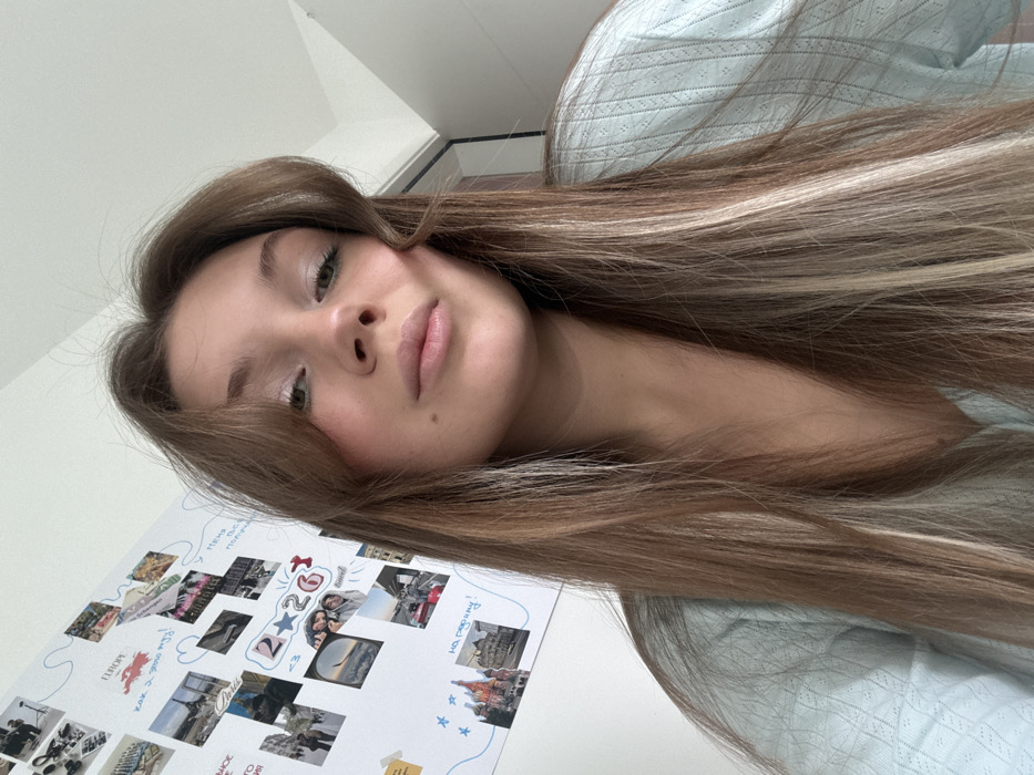 A young female, appearing to be between 18 and 25 years old, is depicted indoors in the city of Vozdovac Urban Municipality, Serbia. She has long, straight to slightly wavy brown hair with blonde highlights, styled with face-framing bangs. Her eyes are green, and a small mole is visible on her left cheek. She is wearing a light blue, V-neck top with a subtle, textured pattern. Her expression is neutral as she looks directly into the camera. The background on the left consists of a white wall adorned with a collage of photographs and handwritten text in Cyrillic script. Visible images include tourist landmarks such as the Eiffel Tower and a cityscape resembling Moscow's Red Square, alongside various candid snapshots of individuals. Text fragments like "EUROPE," "2 26," and "на райони!" are identifiable. A dark vertical element, possibly a door frame or furniture edge, is partially visible on the right side of the frame.