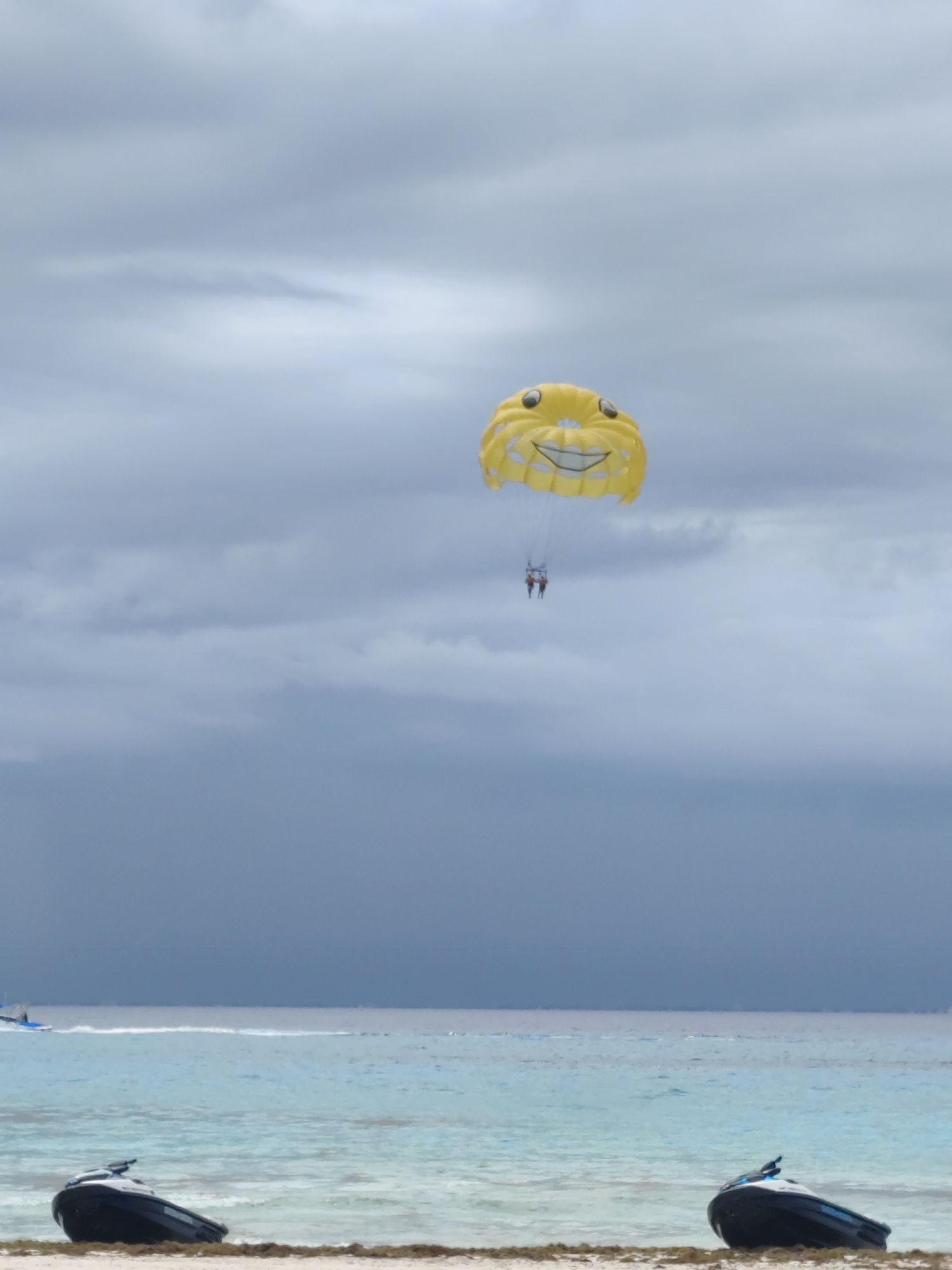 The image shows a beach scene in Playa del Carmen, Mexico. In the foreground, two black jet skis are resting on the sand near the water. The water is a turquoise color. In the distance, the sea meets a gray, cloudy sky. A boat is slightly visible in the water. In the center of the image, a yellow parasail with a smiley face design carries two small figures against the cloudy sky.