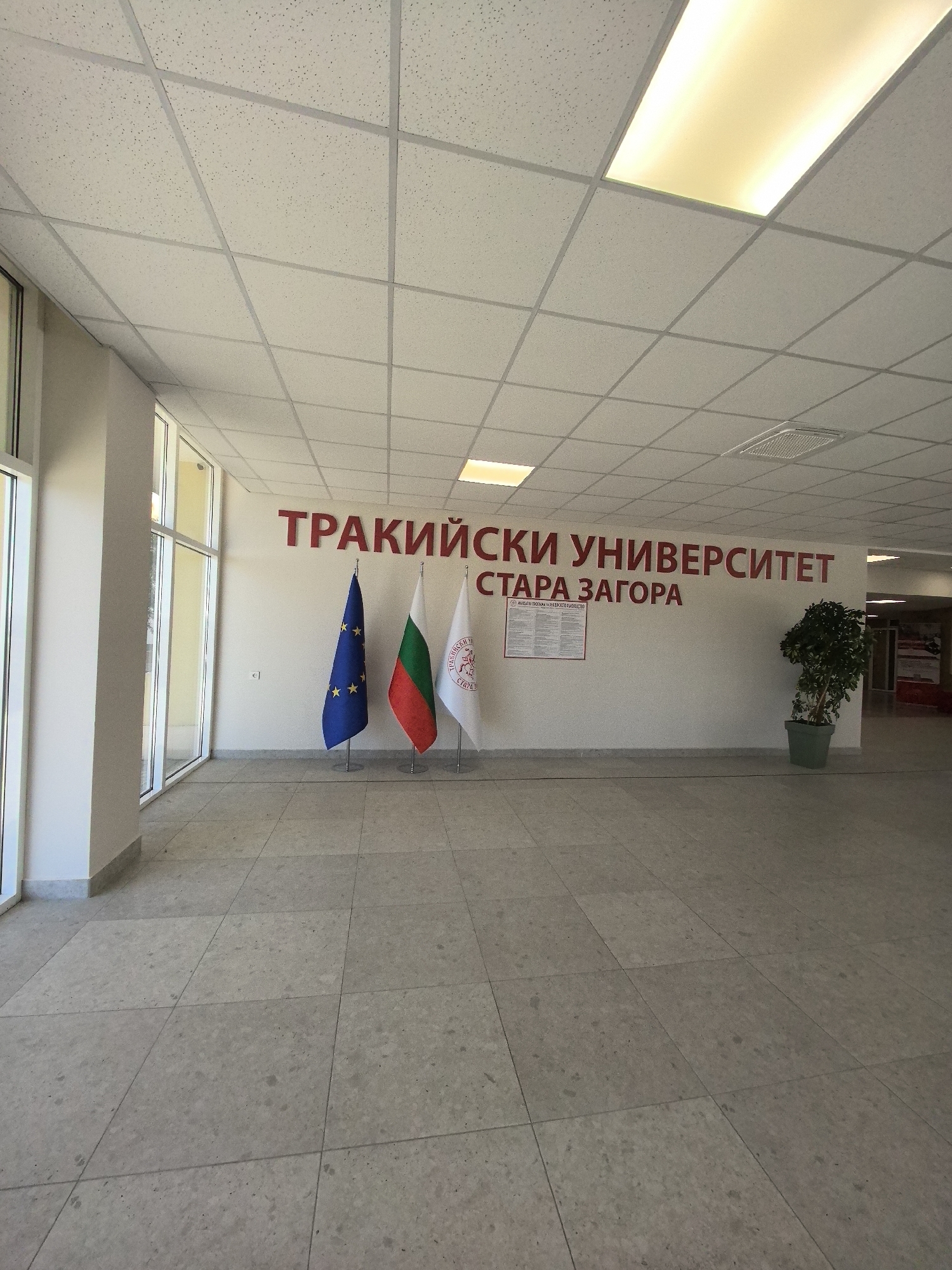 The image depicts the interior of an academic building. The wall is adorned with the inscription "ТРАКИЙСКИ УНИВЕРСИТЕТ СТАРА ЗАГОРА", in red lettering, indicating the location is likely related to the Thracian University of Stara Zagora. Three flags are displayed: the European Union flag, the Bulgarian national flag, and the university's flag. A potted plant is situated near the wall. The floor is tiled, and the ceiling is a suspended grid system with integrated lighting. There are glass windows. The specific city is not identified.