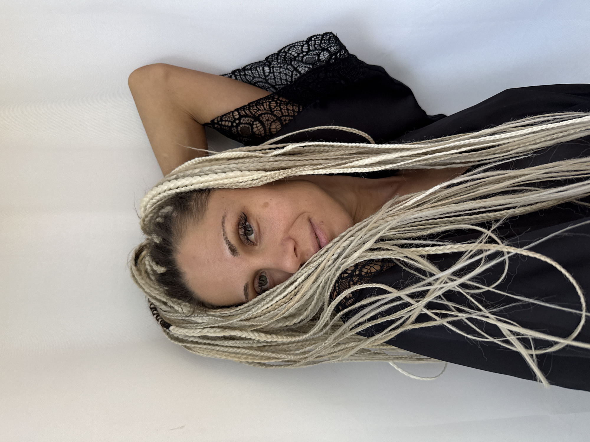 In this medium shot, a woman with blonde braids is positioned against a plain white background. She is looking towards the camera with a slight smile. Her left arm is raised behind her head, and she is wearing a black garment with lace detailing on the sleeves and neckline. The lighting is even, illuminating her face and hair. There is no indication of the location being in Rusanovo, Russia.