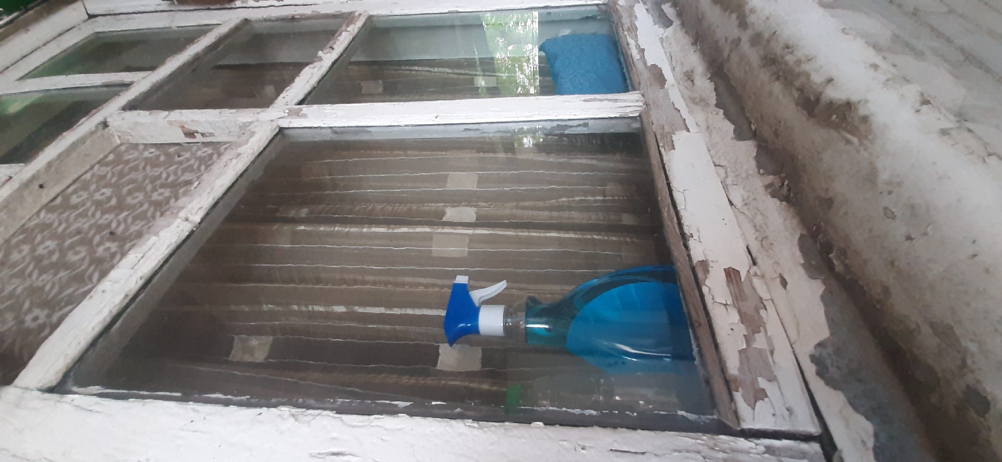 A close-up shot shows a window in Yessentukskaya, Russia, with a spray bottle containing blue liquid and a blue sponge visible on the sill. The window frame is old and painted white, with peeling paint. The glass is dirty, and behind it, vertical blinds or curtains are visible, with a patterned overlay on the upper portion. The sill itself appears worn and stained. The lighting suggests an indoor environment, possibly a residential or older commercial building.