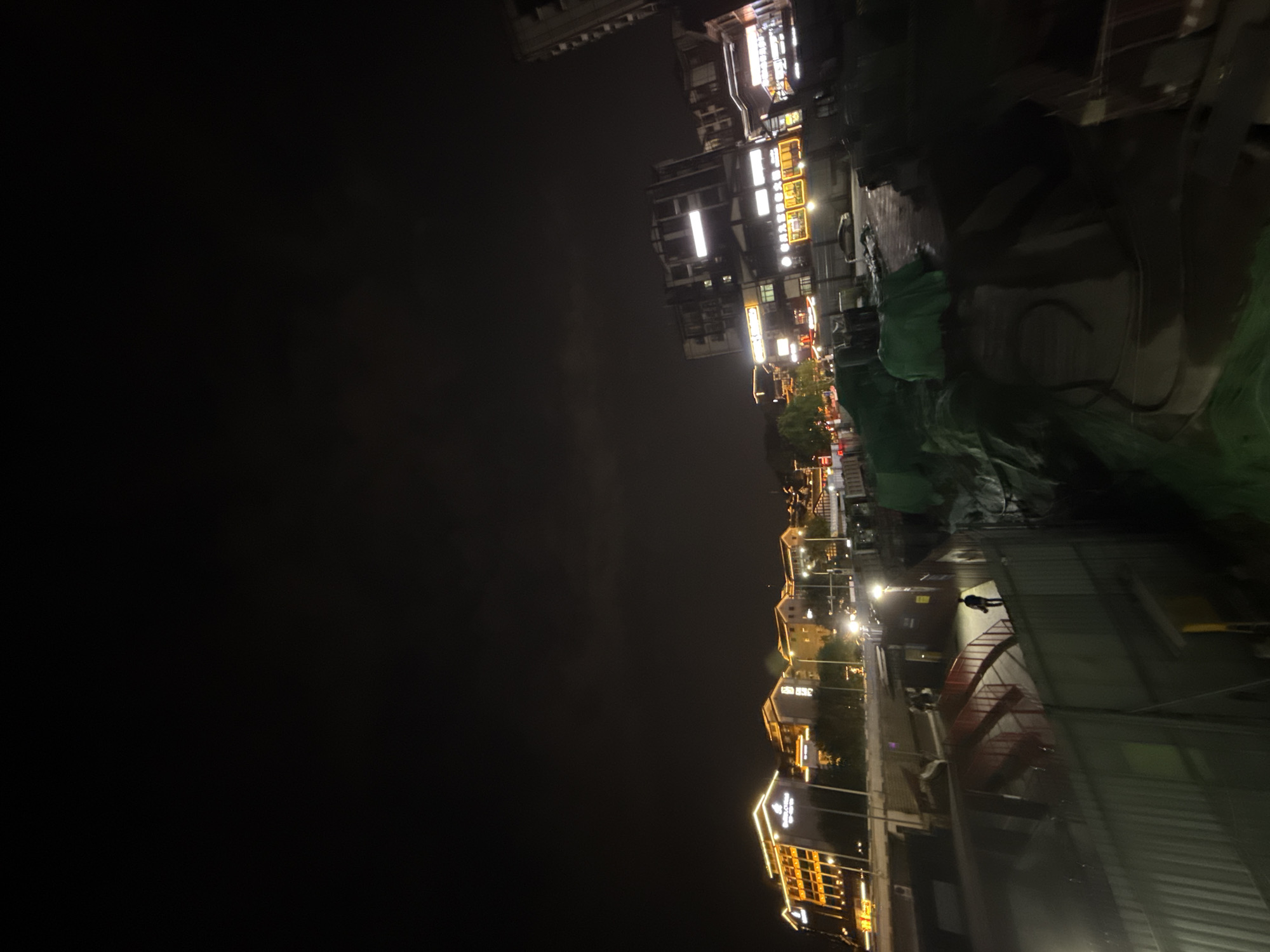 A nighttime photograph captures an illuminated urban landscape, likely a street or riverfront area, under a dark sky. The scene is dominated by artificial lights from buildings and street lamps.
On the right side of the image, multiple multi-story buildings line what appears to be a street or riverbank. These structures are brightly lit, showcasing numerous glowing windows and illuminated commercial signs, some featuring indecipherable Chinese characters. The lights suggest a lively commercial or residential area.
In the lower right foreground, a solitary figure is visible, walking away from the camera on a well-lit pathway. This path is bordered by streetlights, which cast a warm glow. Adjacent to the path, a large section of the ground or a structure is covered with green netting or tarpaulins, possibly indicating a construction site or protected area. A distinct red structure, potentially a set of stairs or a ramp, is also visible near this green covering.
The left half of the image consists primarily of the dark night sky, with a hint of cloud cover or distant light pollution near the horizon line. Reflections of the building lights are visible on what might be a wet surface or a body of water in the lower part of the frame. The overall atmosphere is quiet, despite the many lights, highlighting the transition from daytime activity to the calm of night in an urban setting. This scene is located in Shapingba District, China.