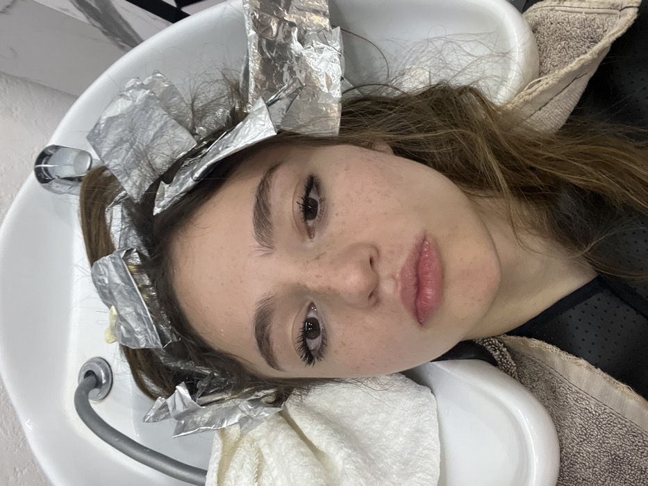 A young female, appearing to be a teenager or young adult, is depicted receiving a hair treatment in a salon located in Puebla, Mexico. Her head is tilted back into a white ceramic hair washing basin, with numerous sections of her brown hair wrapped in silver foil, indicating a chemical process such as highlighting or coloring. A chrome faucet is visible on the right side of the basin, and a grey flexible hose or spray nozzle is on the left. The female has a fair complexion with visible freckles across her nose and cheeks. Her dark eyes are open, and she wears mascara. Her lips are slightly pursed, forming a subtle pout. A white terry cloth towel is draped around her neck, resting on the left rim of the basin, and she wears a dark, possibly black, perforated smock or cape over her shoulders. The immediate environment consists of the salon's washing station.