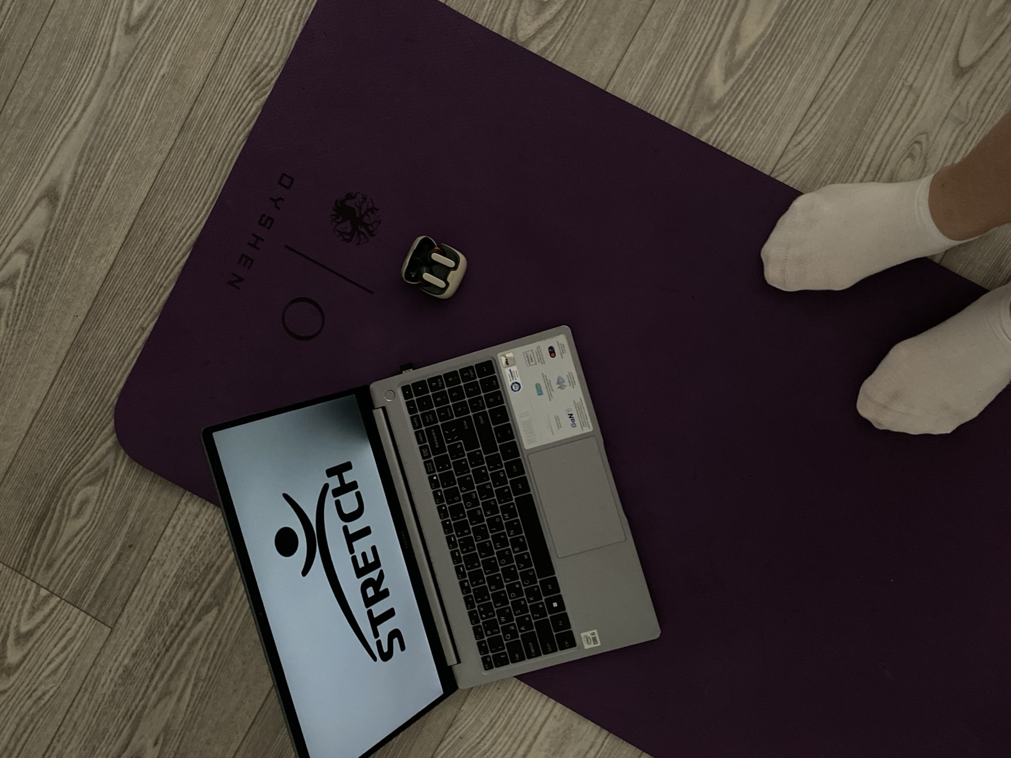 Here is a forensic description of the image.
**Setting:**
The scene is an indoor setting with a light-colored wooden floor. The floor appears to be made of planks and is clean and relatively uncluttered.
**Subjects and Objects:**
1. **Yoga Mat:** A large, purple yoga mat is prominently placed in the center of the frame. It is slightly angled. The brand name "DYSHEN" is printed along one edge of the mat.
2. **Laptop:** A laptop is positioned on the yoga mat. The screen is open, displaying a logo with the word "STRETCH". The keyboard is visible and appears to be a Russian/English layout.
3. **Earbuds Case:** A small, white earbuds case is located on the yoga mat near the top left of the keyboard.
4. **Human Presence:** Two feet wearing white socks are visible on the top right of the yoga mat. The person is likely standing or positioned beside the mat.
**Action:**
The image depicts a setup for a stretching or yoga session, with a laptop potentially providing instructions or entertainment. The person is ready to participate, as indicated by their presence near the mat.
**Environment:**
The scene is simple and minimalistic, focusing on the yoga activity.
Based on the keyboard layout, the location could be in Russia. Therefore, the setting could be in Sochi, Russia, but this cannot be determined with certainty based solely on the image.