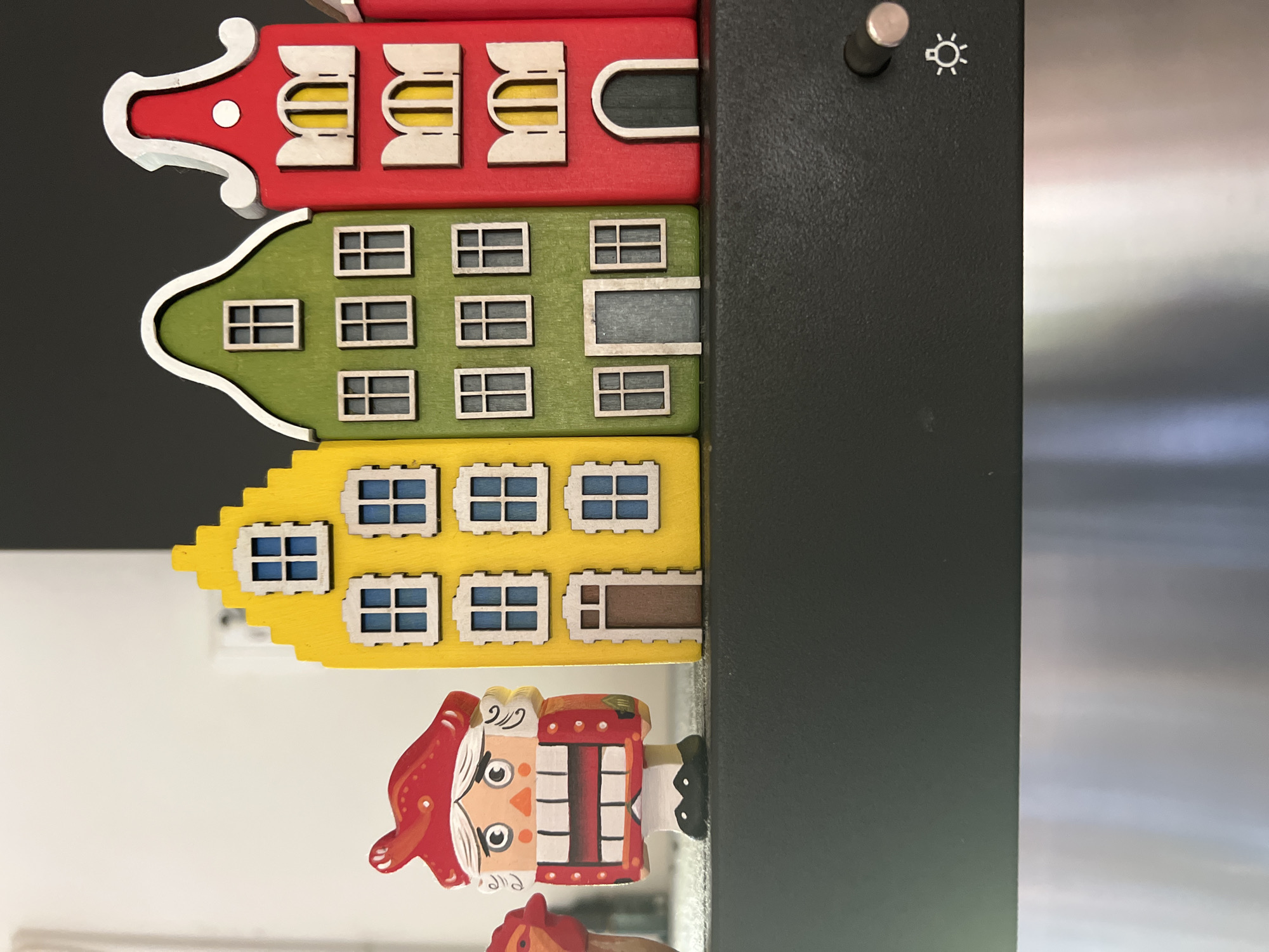 A close-up, eye-level shot shows a collection of decorative wooden Dutch houses and a nutcracker displayed on a dark gray surface, possibly a shelf or countertop. The houses are painted in vibrant colors: yellow, lime green, and red. They are designed with stepped gables and feature white-framed windows and doors. The nutcracker, positioned to the left of the houses, is a traditional wooden figure painted in red and white with black accents, a red hat, and a stern expression. The background consists of a white wall on the left and a dark gray surface on the right. The lower portion of the image shows a metallic, reflective surface. A small circular silver knob and a lightbulb icon are visible on the dark gray surface in the lower right corner. The scene appears to be indoors, in an unknown urban setting.