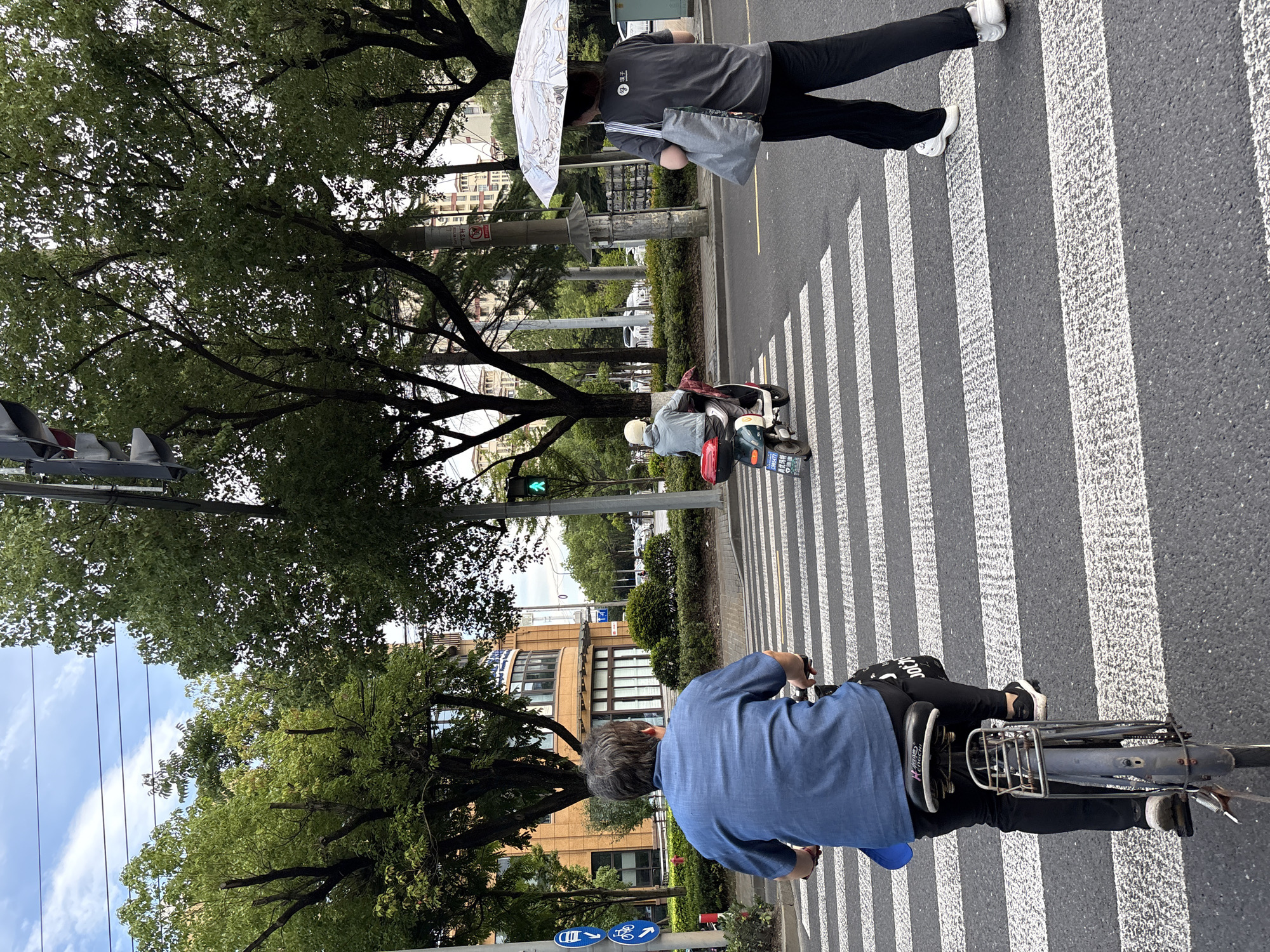 In Minhang District, China, on a clear day, three individuals are captured crossing a pedestrian crosswalk. In the foreground, a person on a bicycle, viewed from behind, is clad in a blue collared shirt and black pants. To the right, another individual walks with an umbrella shielding them from the sun. They are wearing a dark t-shirt and black pants. In the middle ground, a person on a scooter, wearing a light-colored helmet and jacket, is also crossing the street. The surrounding environment features lush green trees, buildings, and traffic signals, indicating a typical urban street scene.